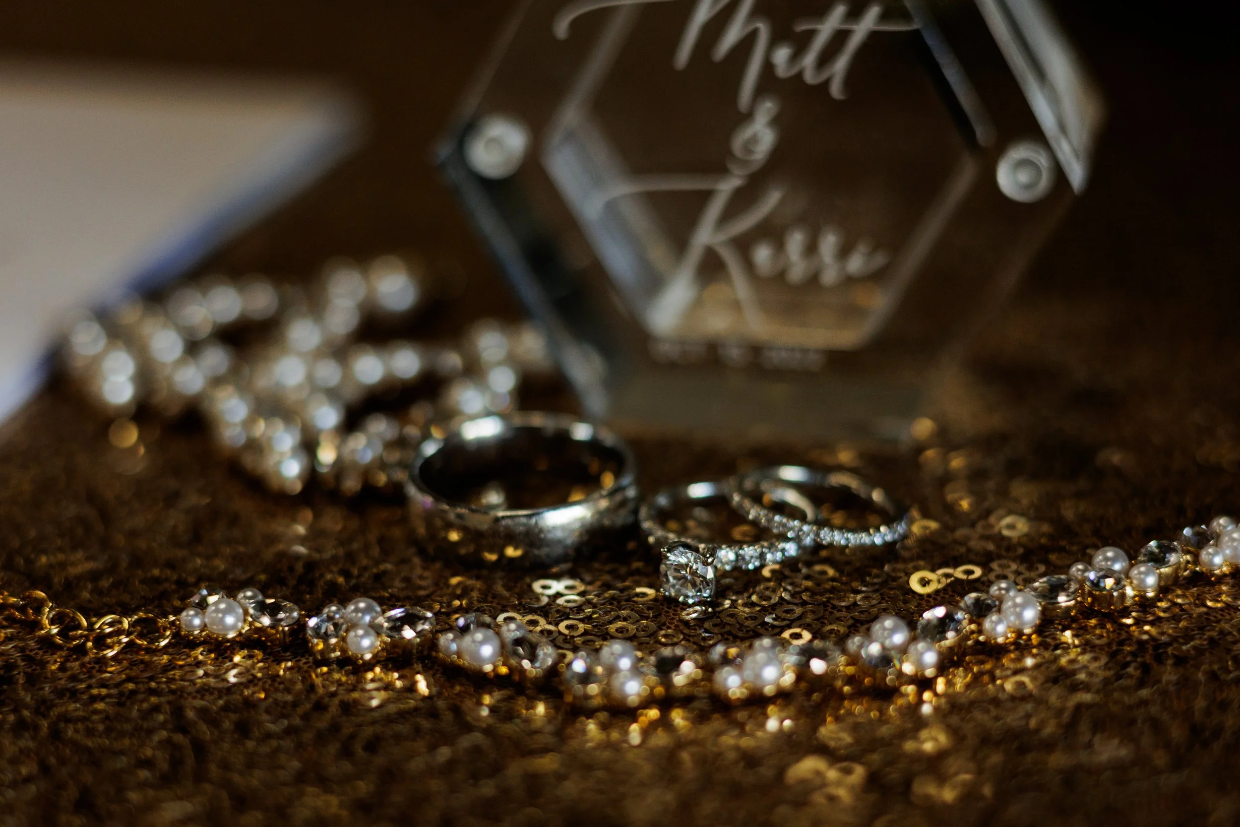 Close-up of wedding rings and jewelry on a glittery surface with a glass decoration that says "Matt & Kassi" in the background.