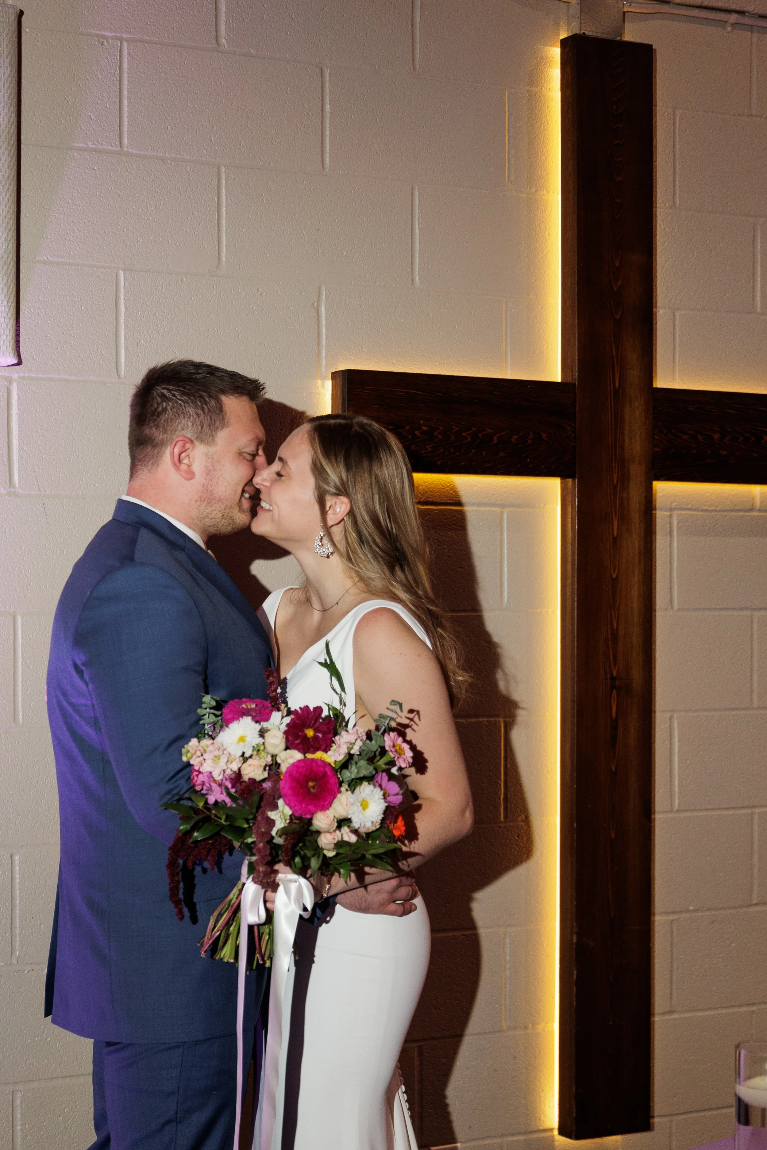 A bride and groom share a kiss in front of a large wooden cross with backlighting, during their wedding ceremony.