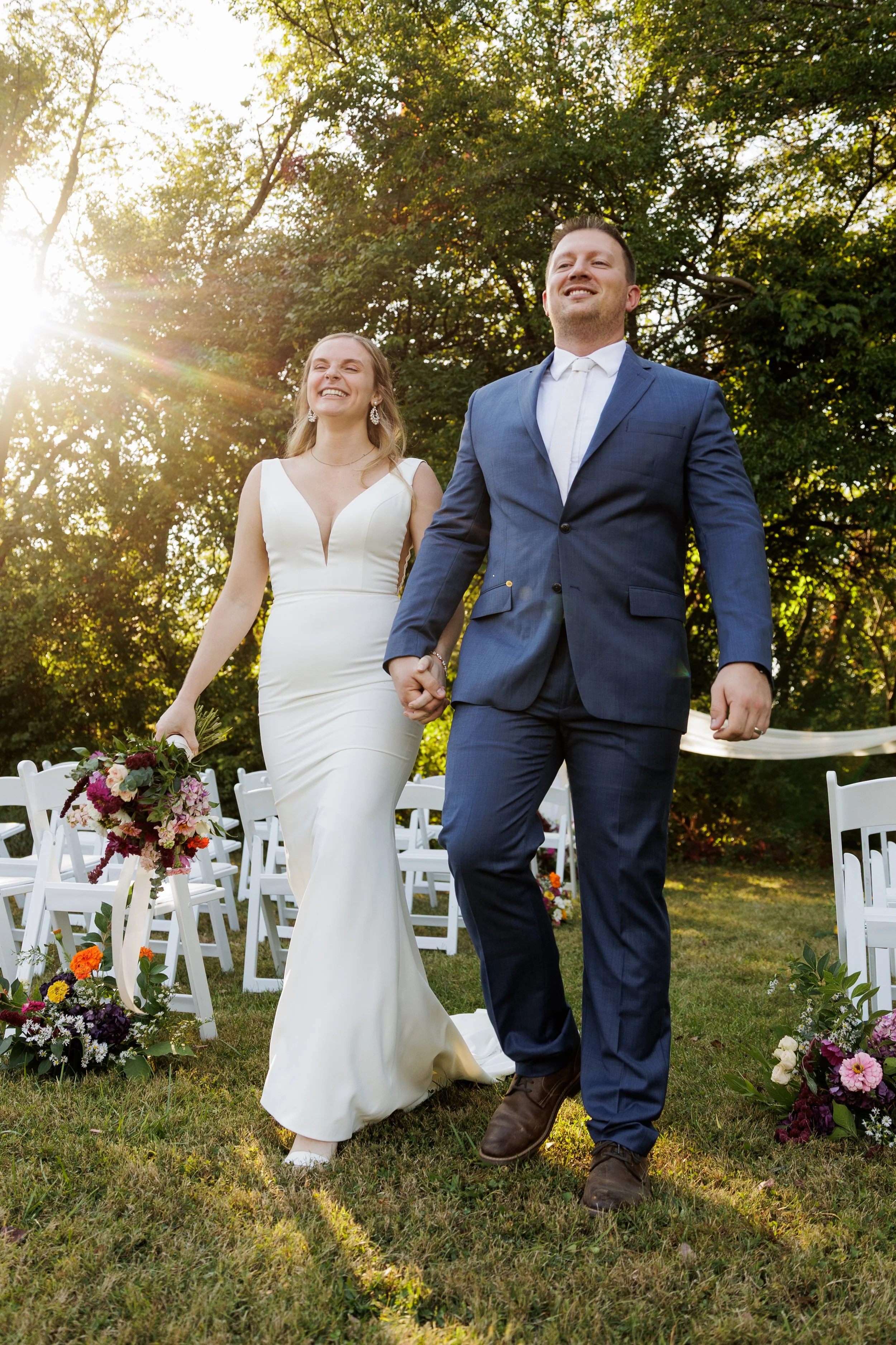 Newlywed couple walking hand in hand outdoors after their wedding ceremony, with chairs and floral decorations in the background and sun shining through trees.