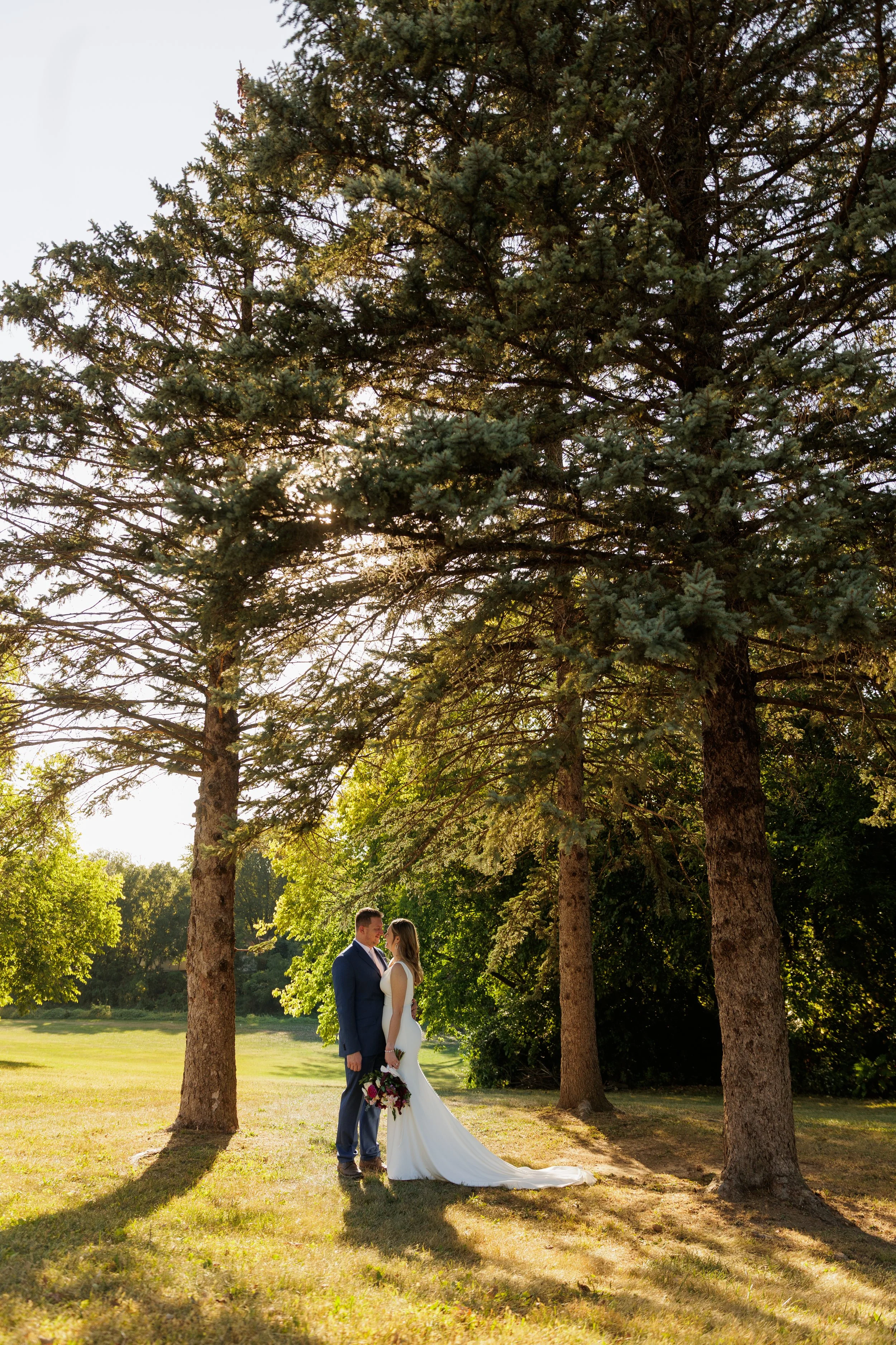 A bride and groom stand facing each other outdoors in a grassy area surrounded by tall trees on a sunny day. The bride holds a bouquet and wears a white wedding gown, while the groom wears a blue suit. They are about to kiss.