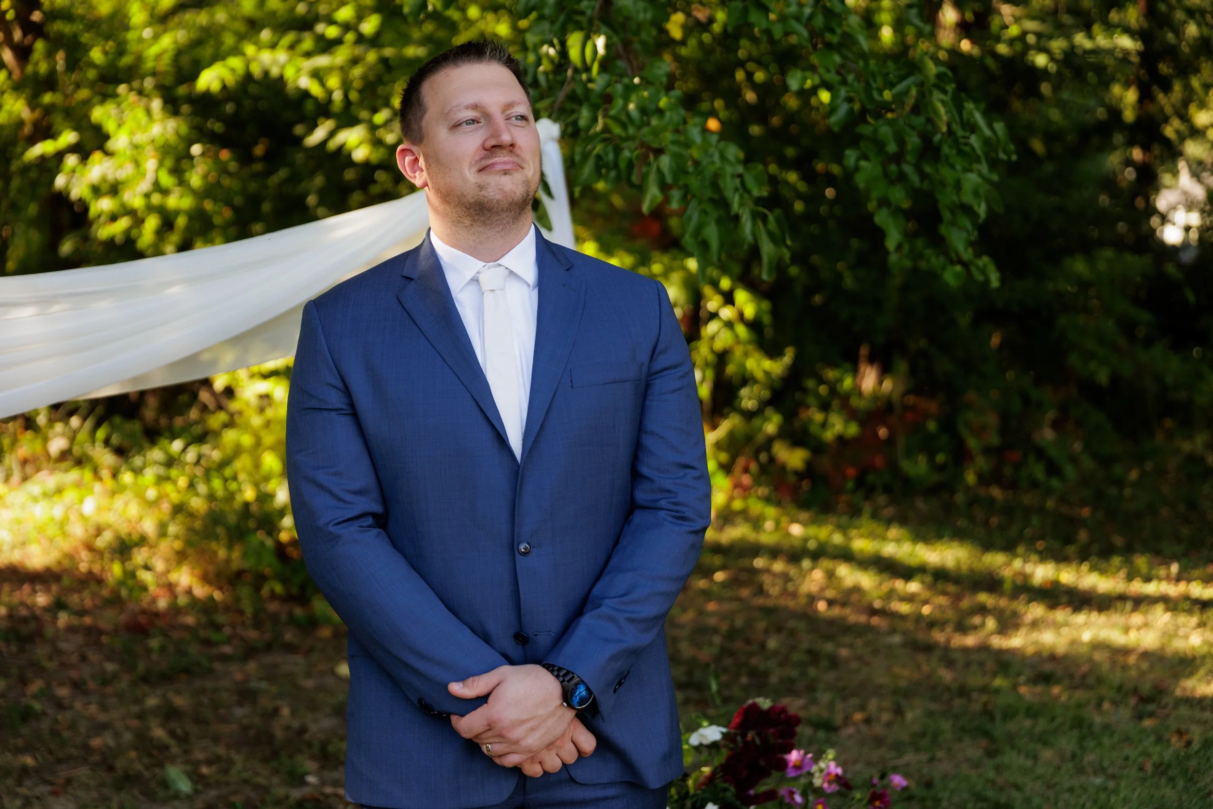 A man in a blue suit stands outdoors, with a serene expression, hands clasped in front of him, surrounded by green foliage and flowers, possibly during a wedding ceremony.