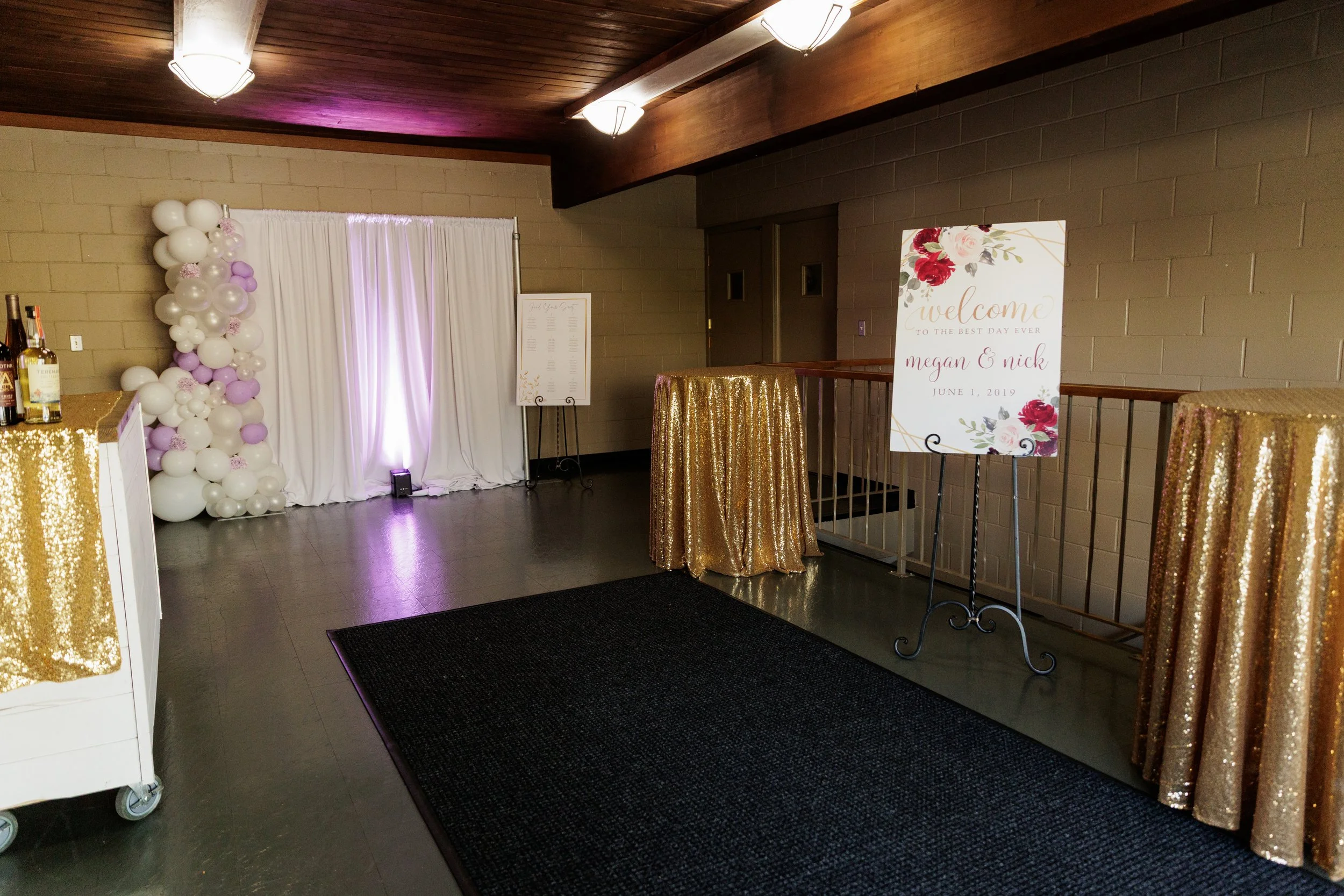 Decorative wedding reception area with a backdrop of white drapes, pink and white balloons, gold sequined tables, and a welcome sign for Megan and Nick, dated June 1, 2019.