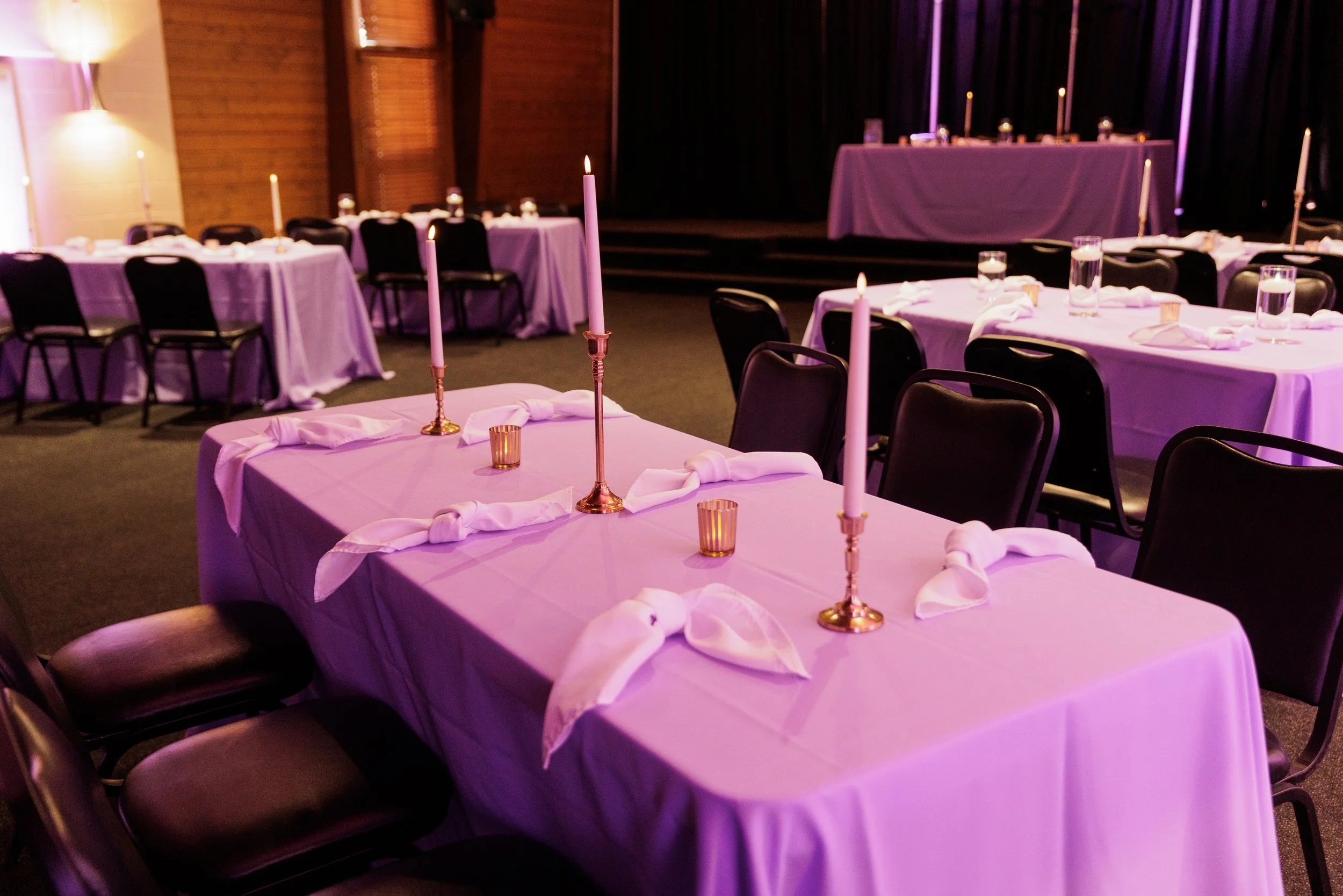 A banquet hall decorated for an event with tables covered in pink tablecloths, adorned with pink candles in gold candlesticks, glasses of water, and napkins tied with ribbons. The room has dark curtains and a stage in the background.