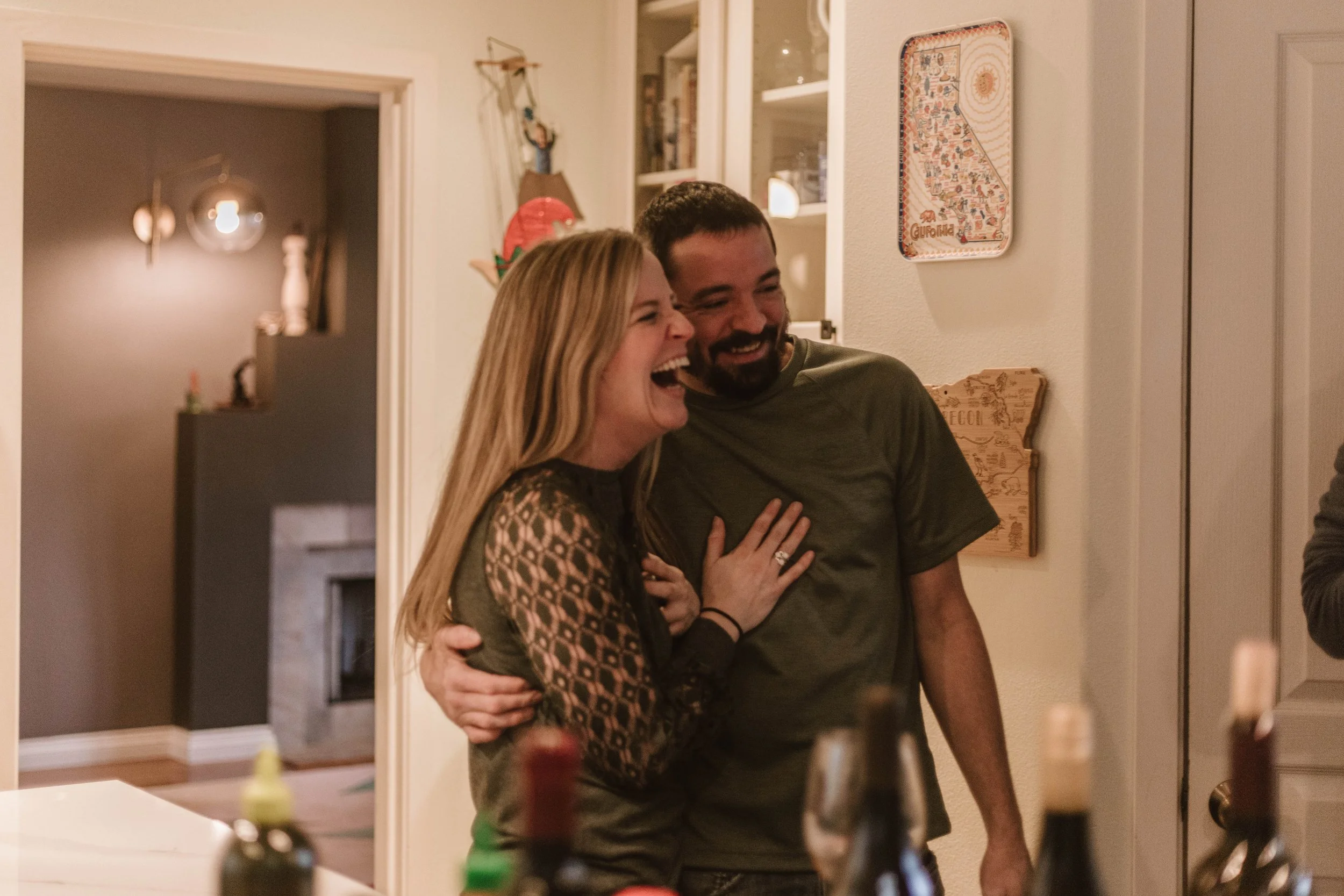 A man and woman laughing and hugging in a cozy indoor setting, possibly a home, with warm lighting and various decorative items on the walls and tables.