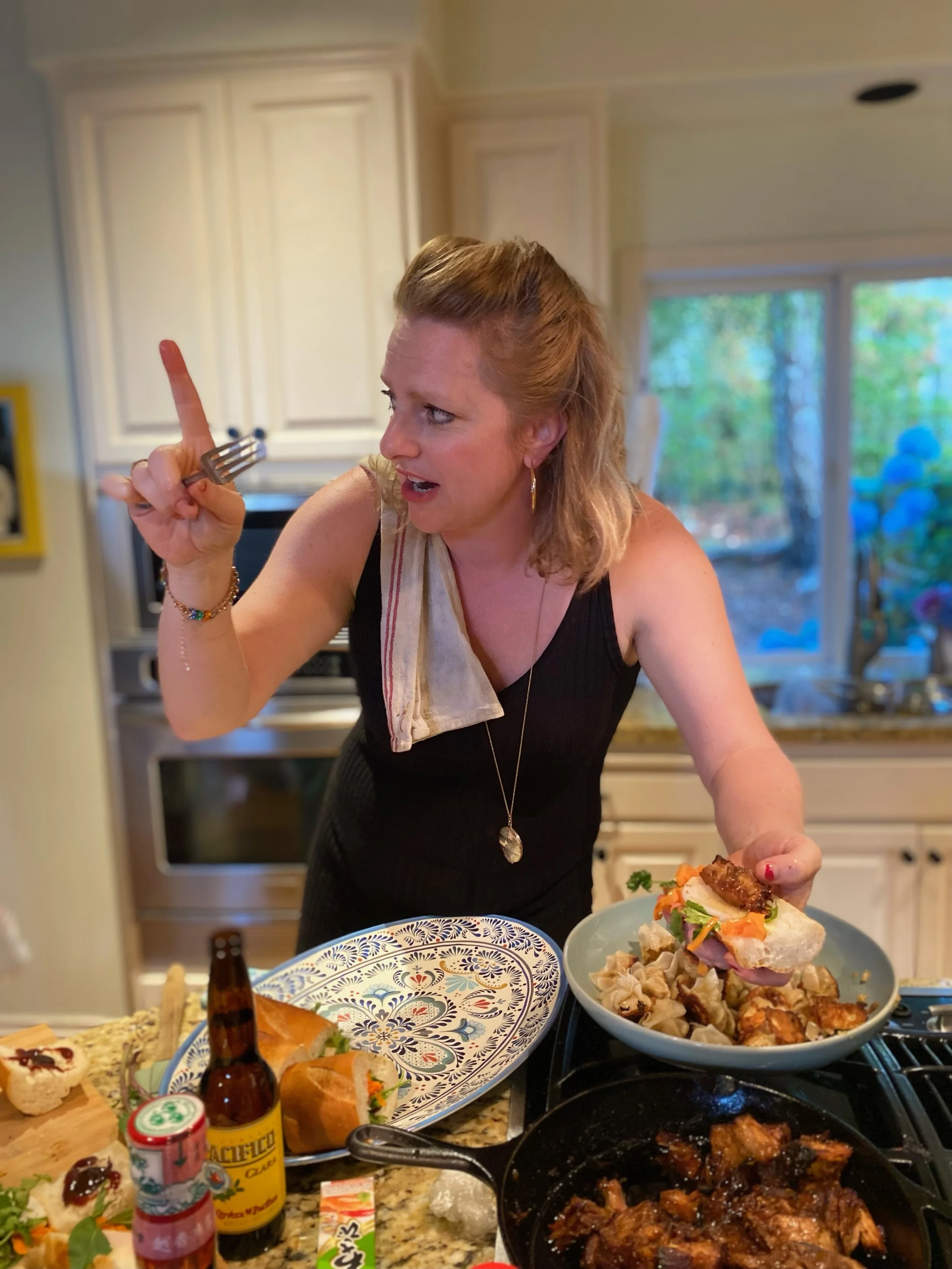 A woman with shoulder-length hair, wearing a black dress and a scarf, is in a kitchen preparing food. She is holding a piece of food in her hand and appears to be engaged in a conversation or explaining something. The kitchen has white cabinets and a large window with greenery outside. There are various dishes and ingredients on the counter, including a bowl of salad, a plate of sushi, bottles of condiments, and a frying pan with cooked meat.
