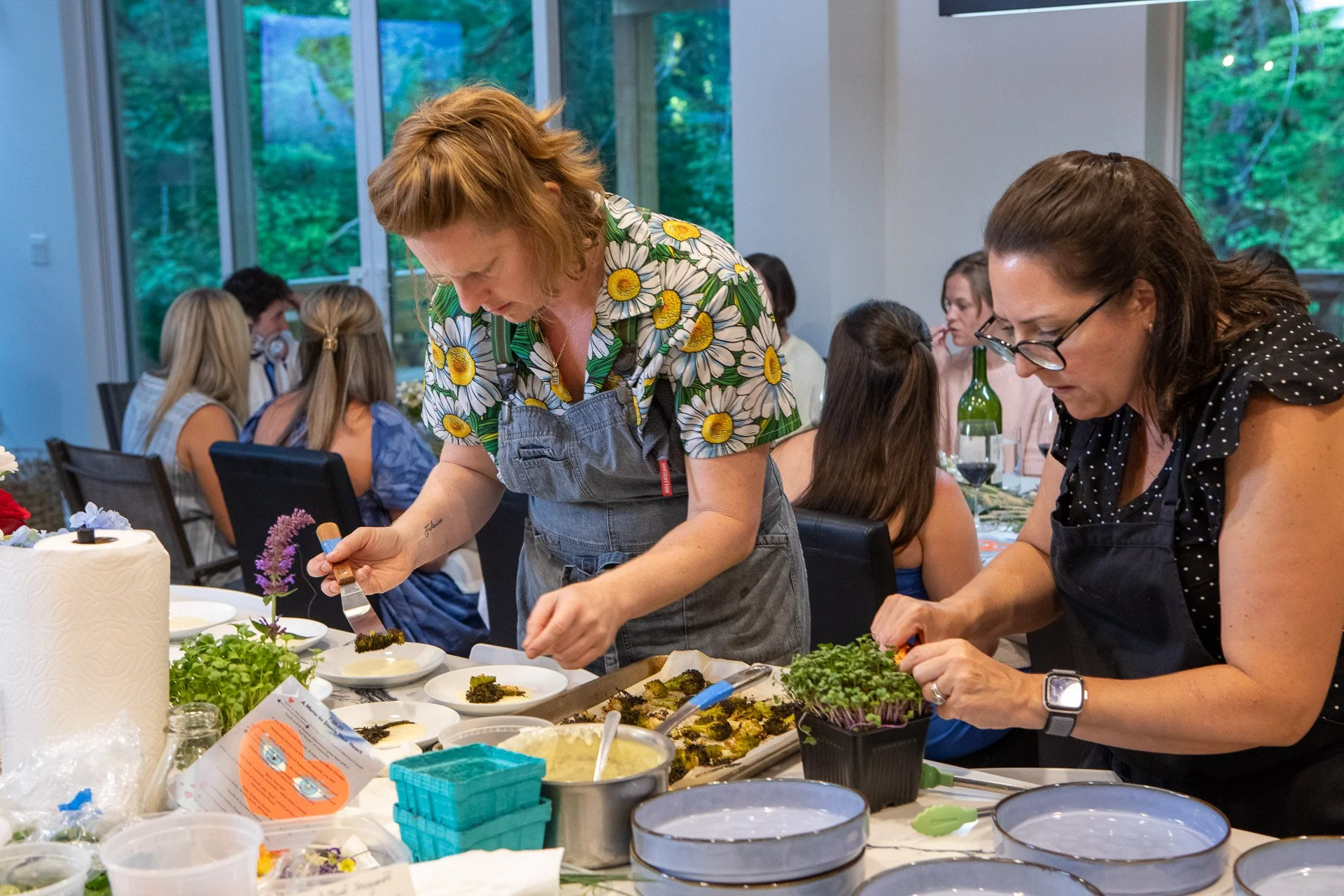 Two women wearing aprons preparing or decorating food at a table with various ingredients and utensils, with a group of people seated in the background inside a room with large windows showing trees outside.