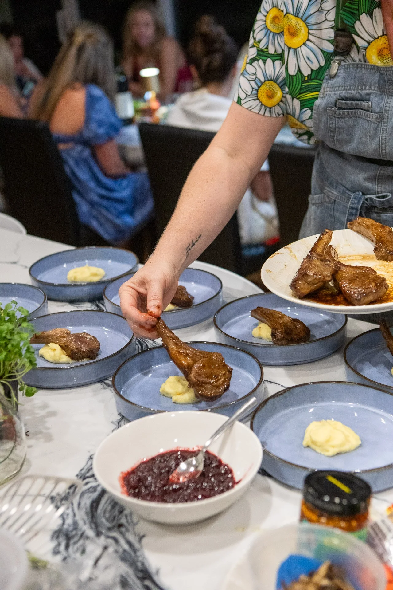Person serving lamb chops with mashed potatoes onto blue plates at a gathering, with other guests in the background.