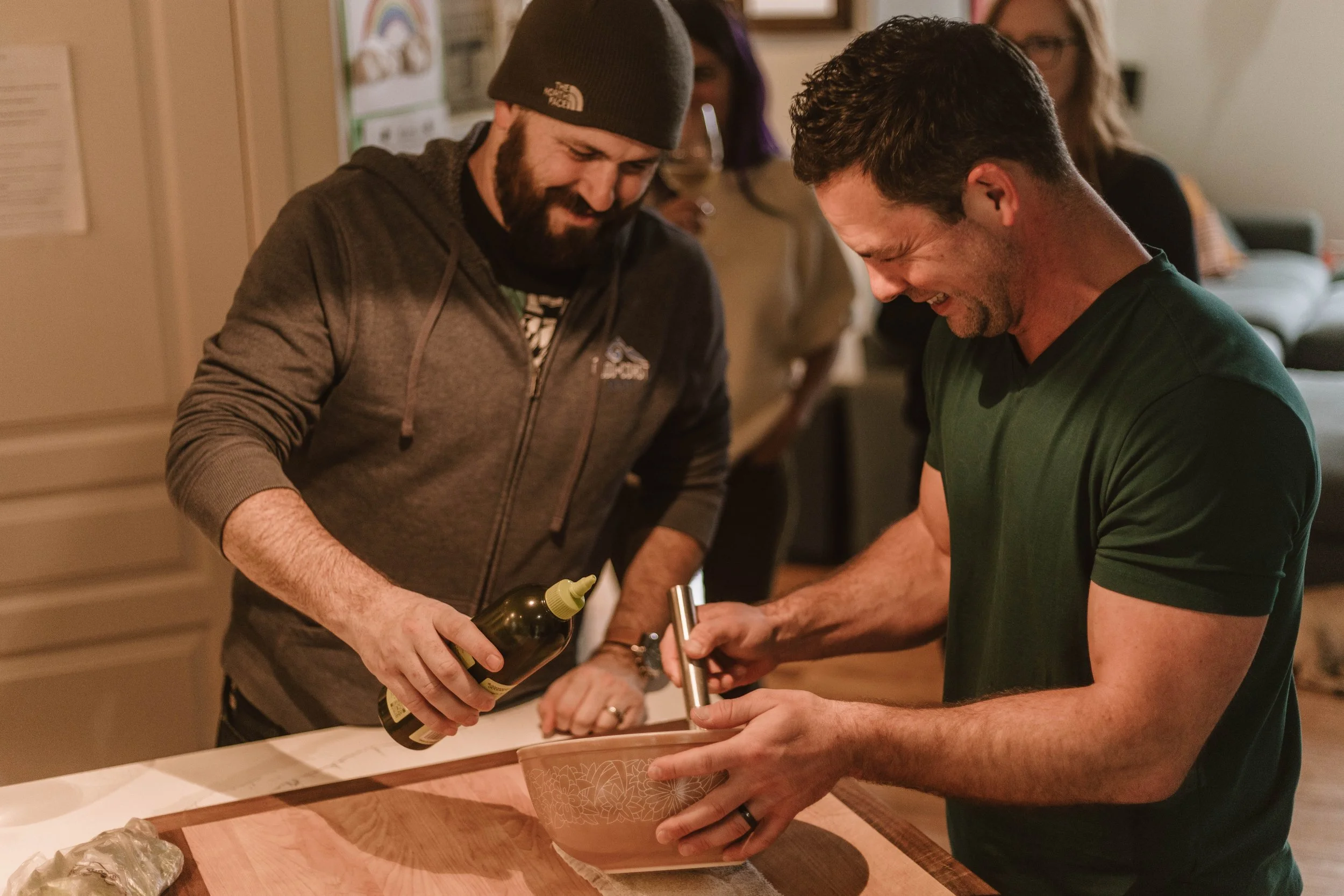 Two men smiling and working together in a kitchen, one pouring a liquid into a mixing bowl, while the other assists. Several people are in the background, and the scene is cozy and well-lit.