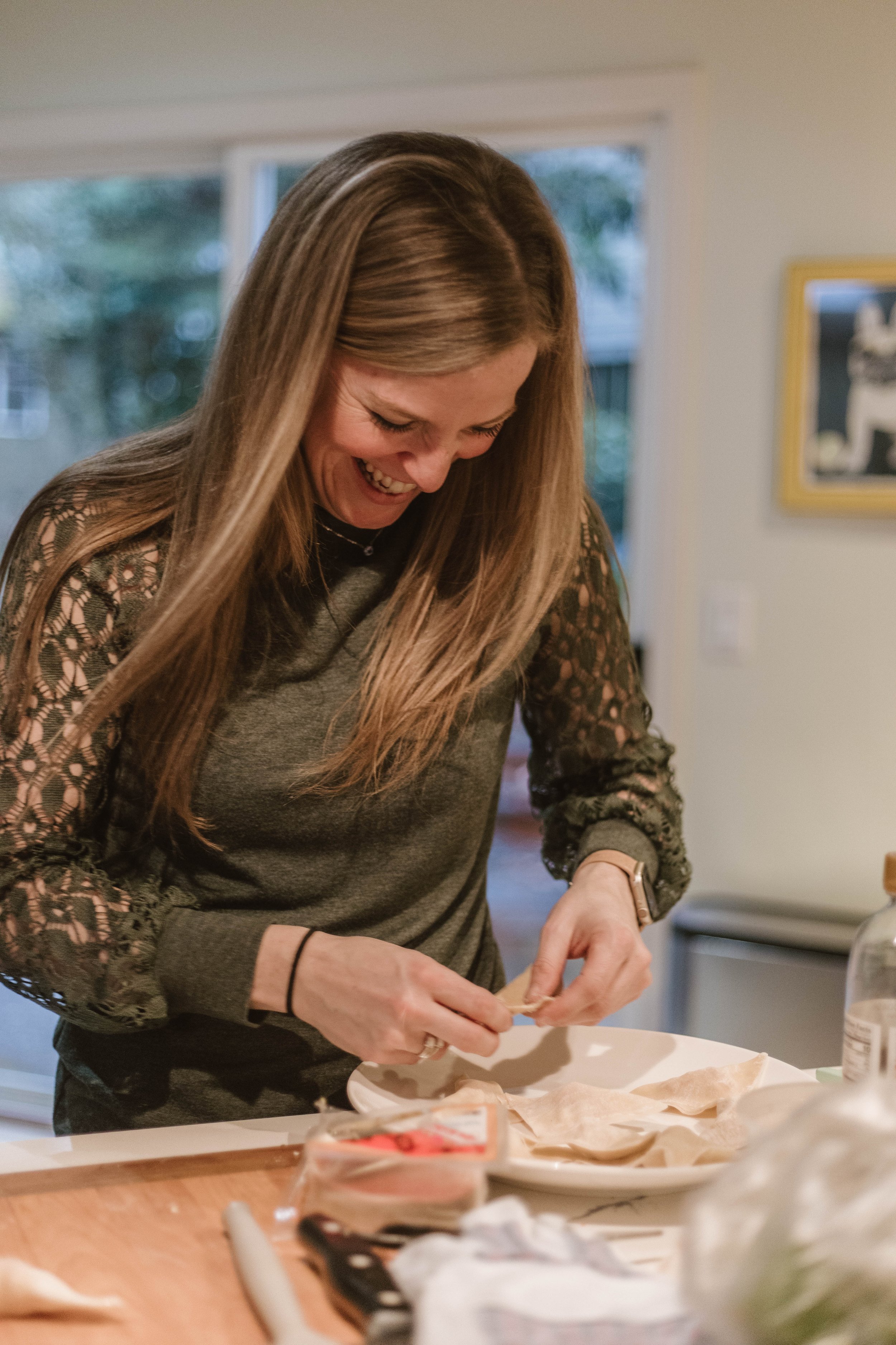 Woman with long hair and lace sleeves peeling skin off a chicken at a kitchen counter surrounded by baking ingredients.