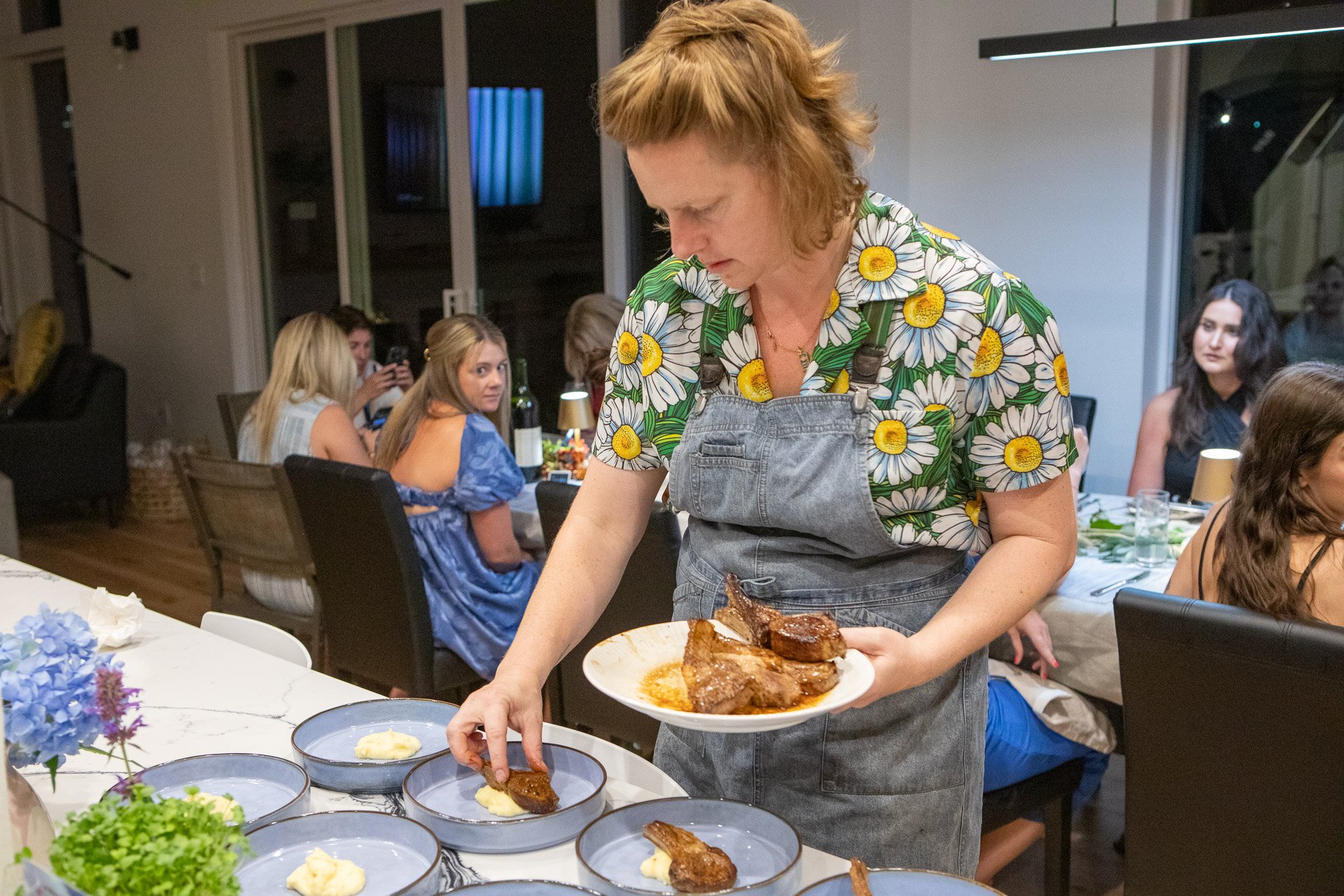 A woman wearing a shirt with daisies and an apron serving food at a dinner party. In the background, guests are sitting at a table, some with phones in hand. The setting appears to be indoors with large windows and a modern interior.