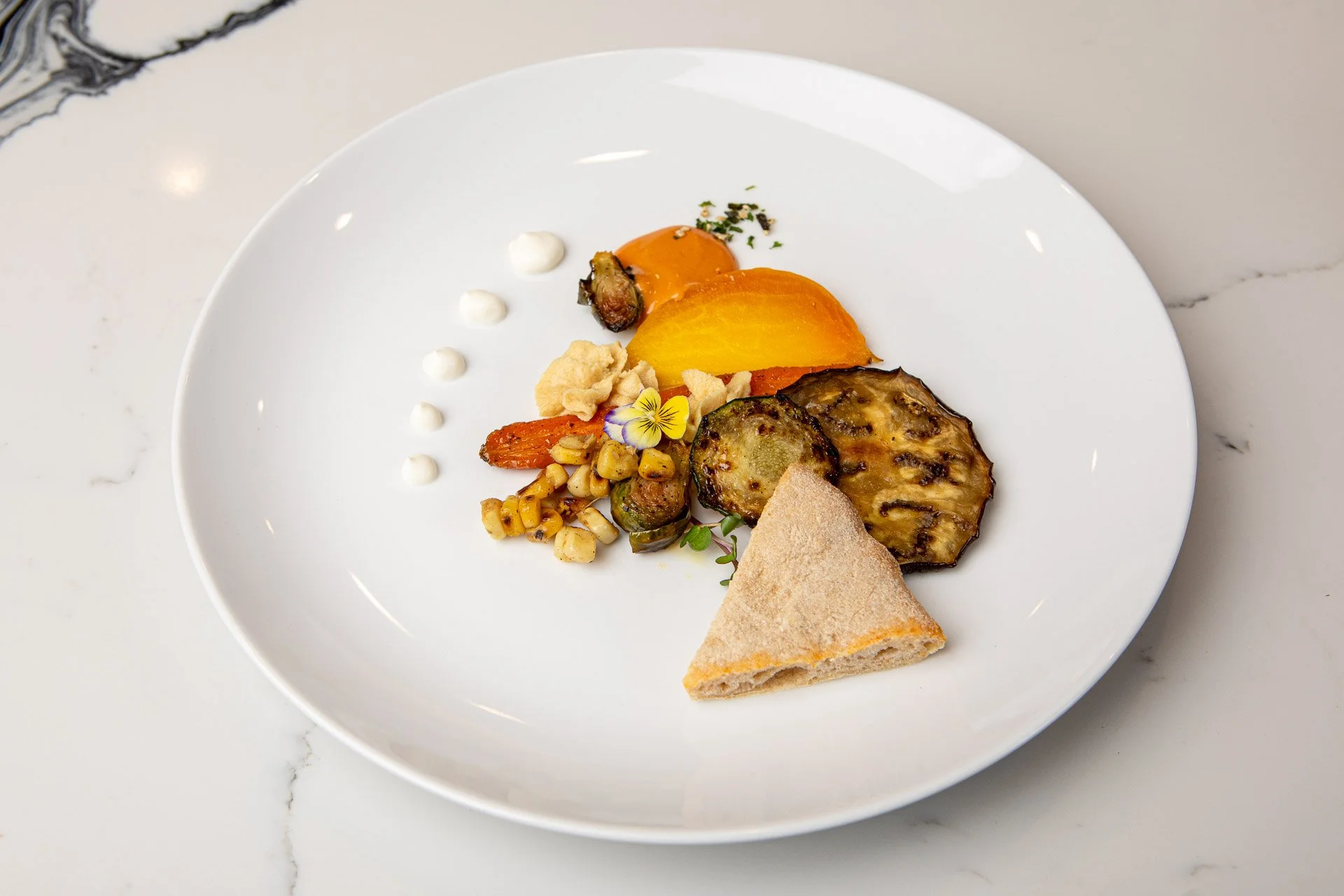 A white plate with assorted grilled vegetables, a piece of flatbread, and decorative dots of sauce, garnished with a small purple flower on a white marble surface.