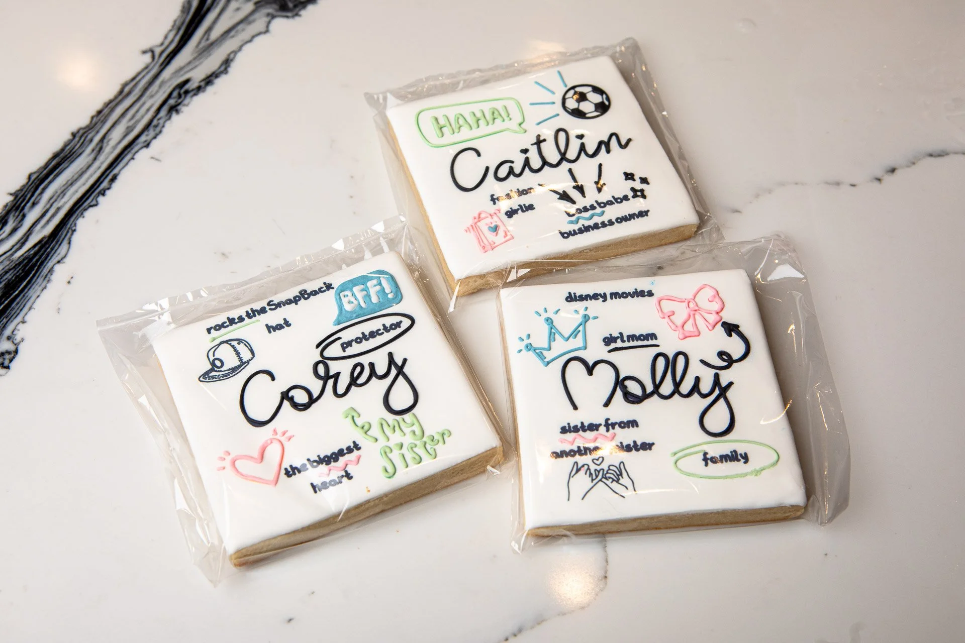 Decorative cookies with personalized messages and illustrations, wrapped in plastic, on a white marble surface.