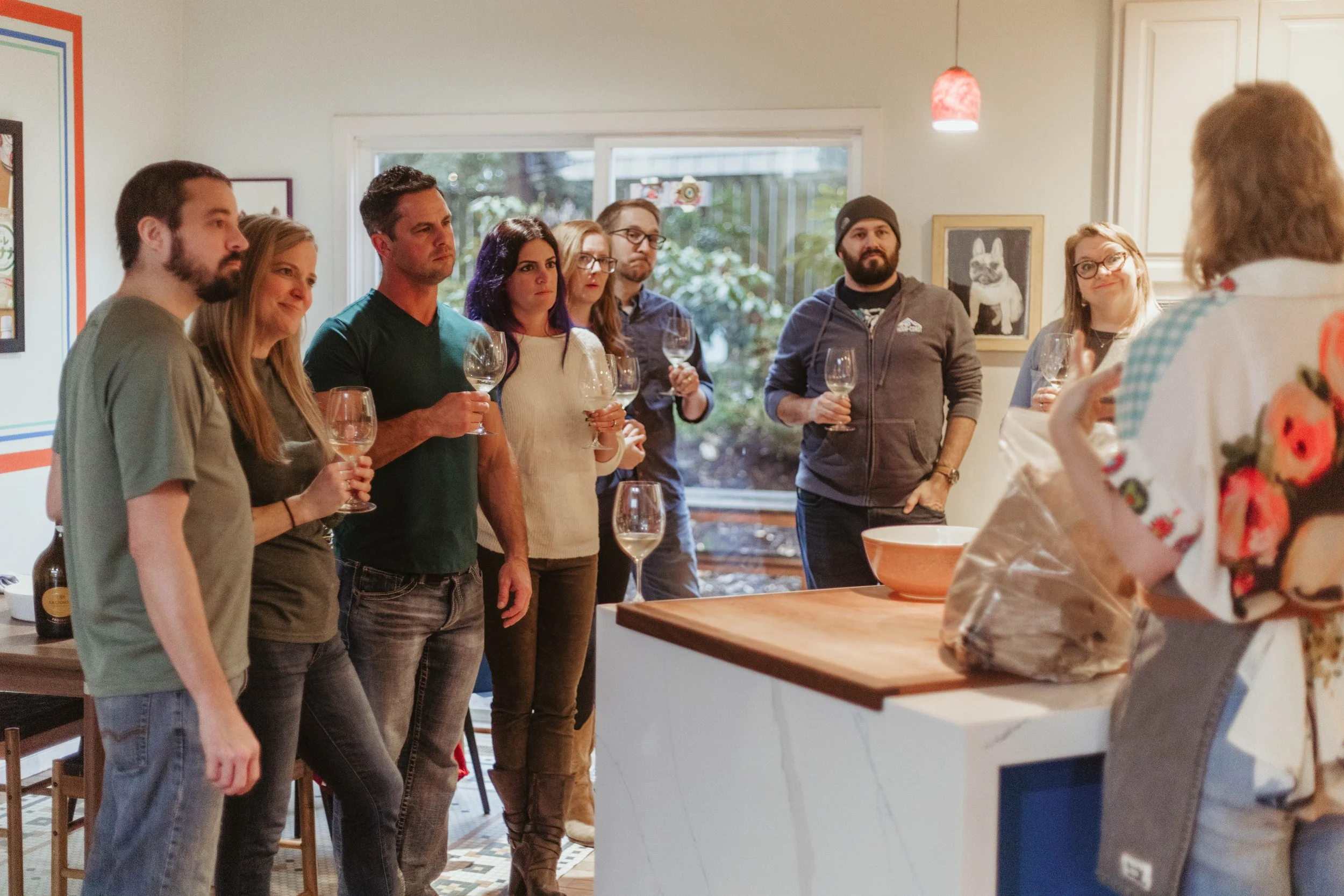 A group of people standing in a kitchen holding wine glasses, listening to a person out of frame. The background shows a window with trees outside and a wall with framed pictures, including one of a dog.