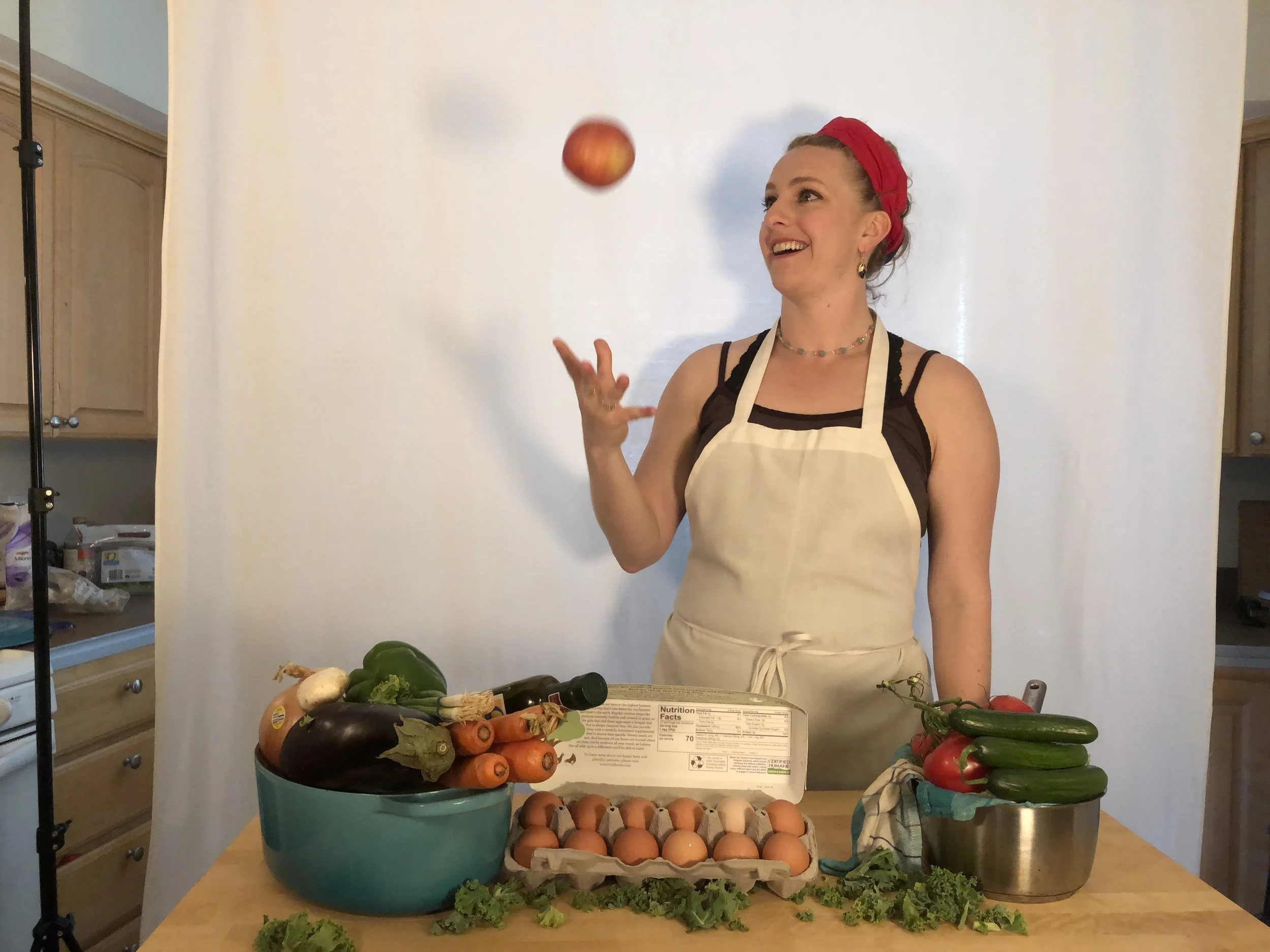 Woman smiling and juggling an apple, standing behind a table filled with vegetables, eggs, and ingredients, with a white backdrop in a kitchen or studio setting.