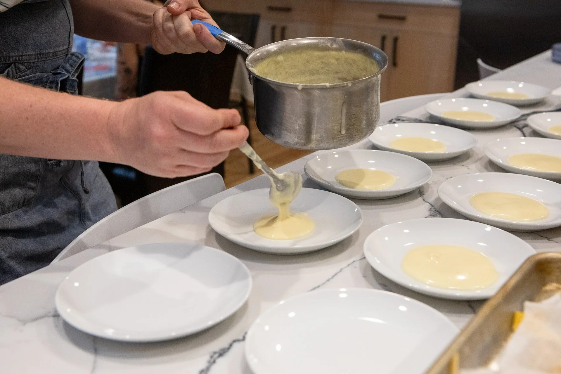 A person is pouring batter into small white bowls, possibly for baking or cooking, with multiple bowls arranged on a white marble countertop.