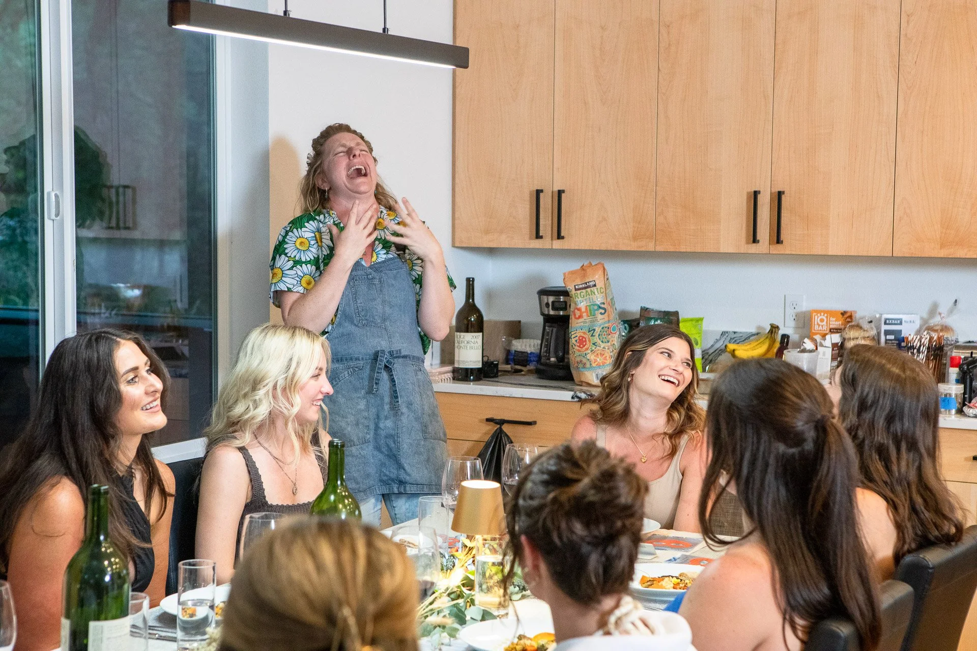 A woman in a floral shirt and denim apron is laughing or crying with her head tilted back, surrounded by other women sitting at a dinner table, in a bright kitchen with wooden cabinets and a tiled backsplash.