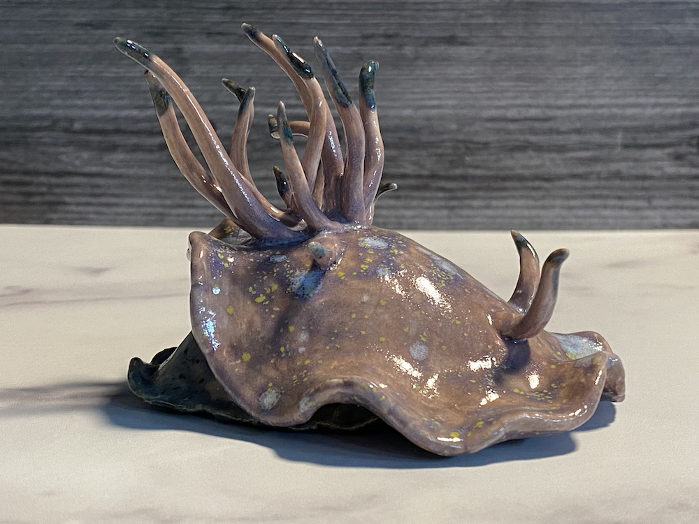Ceramic sculpture of a squid with elongated tentacles, glossy finish, and a speckled pattern, placed on a light-colored surface.