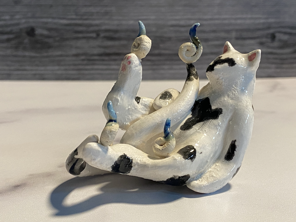 Ceramic sculpture of a cat with curled tail and four small kittens, painted with black and blue accents, on a white surface with a gray wooden background.