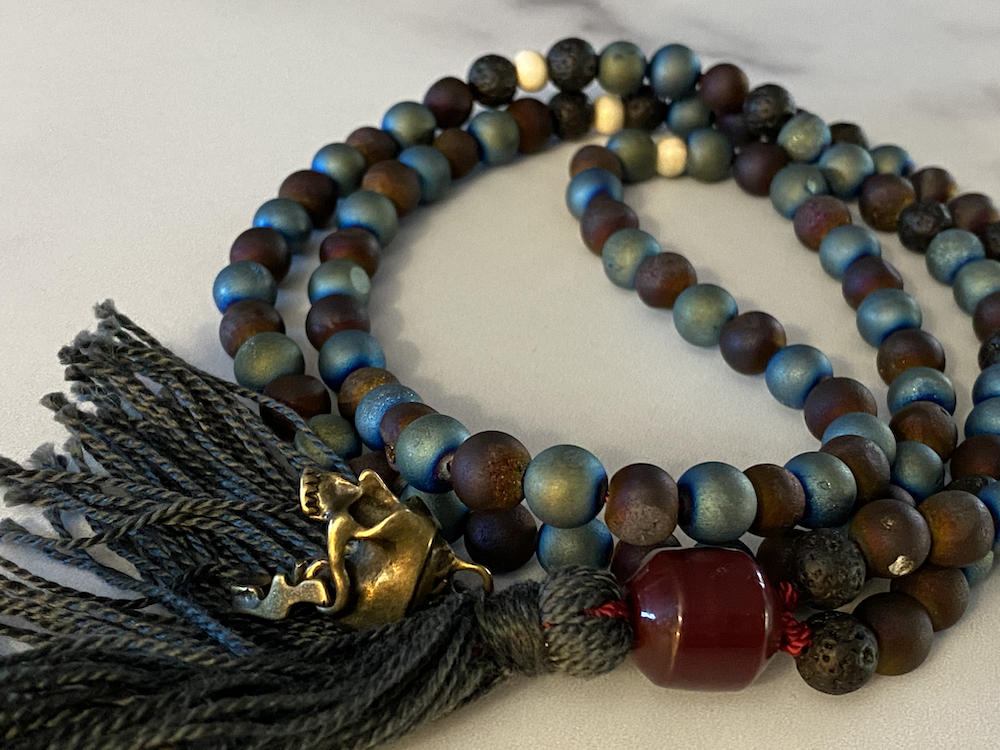 Beaded necklace with multicolored matte beads, a large red bead, a gold-tone decorative element with a black tassel, and gray tassels at the end, arranged on a white surface.