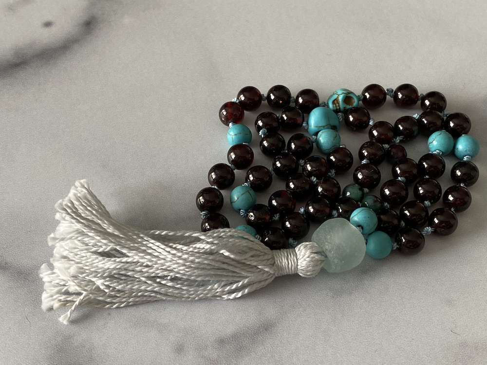 Close-up of a beaded necklace with turquoise and dark red beads, white tassel at one end, lying on a smooth, gray surface.