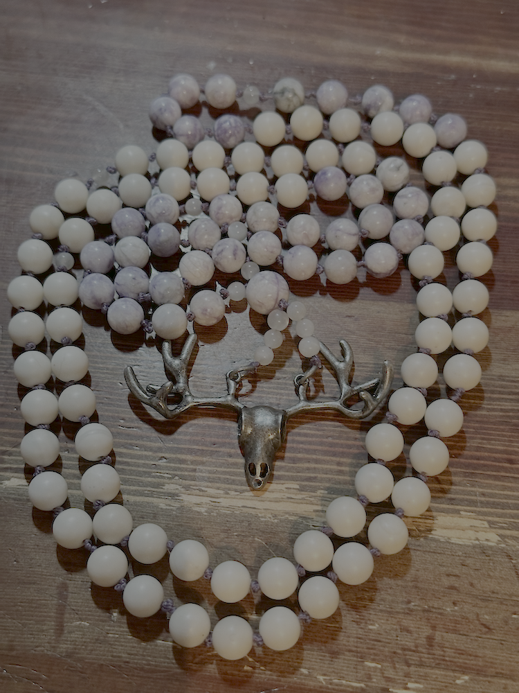 A necklace made of white and purple marbled beads with a silver deer skull pendant, arranged in a circle on a wooden surface.