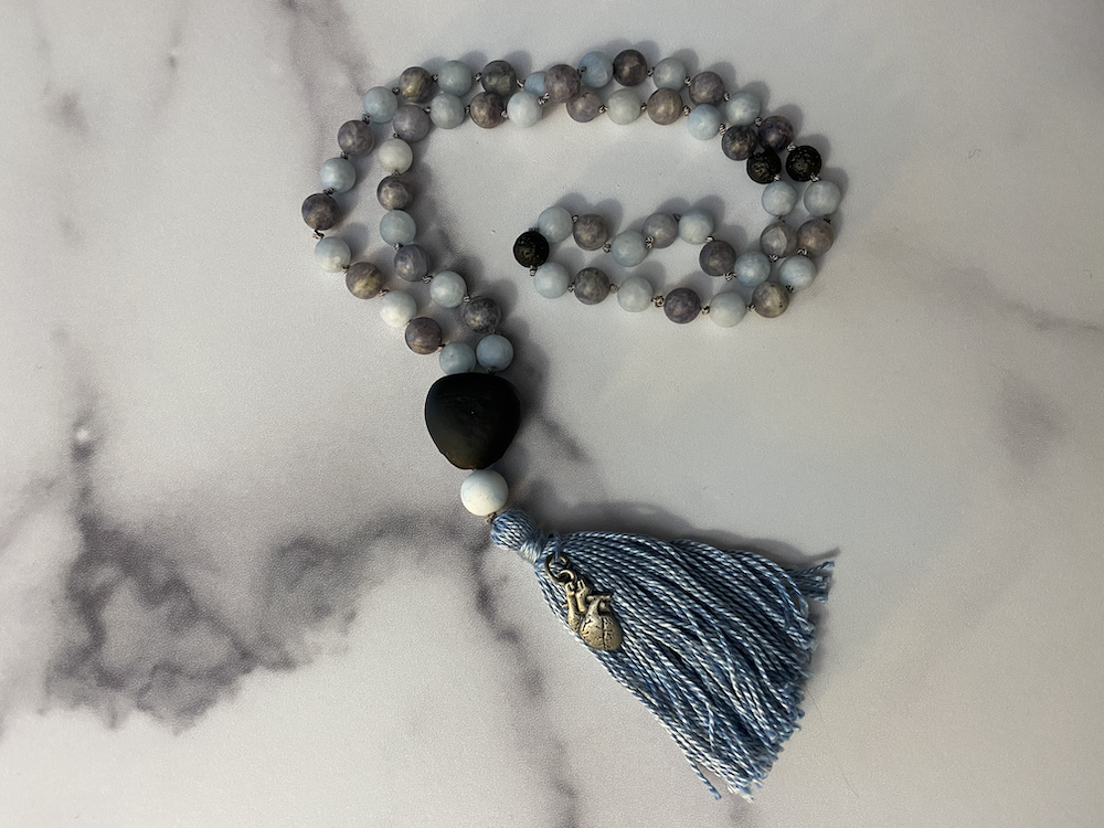 A beaded rosary with white, gray, and black beads, a large black bead, a smaller white bead, a decorative silver bead, and a blue tassel at the end, arranged on a light surface.