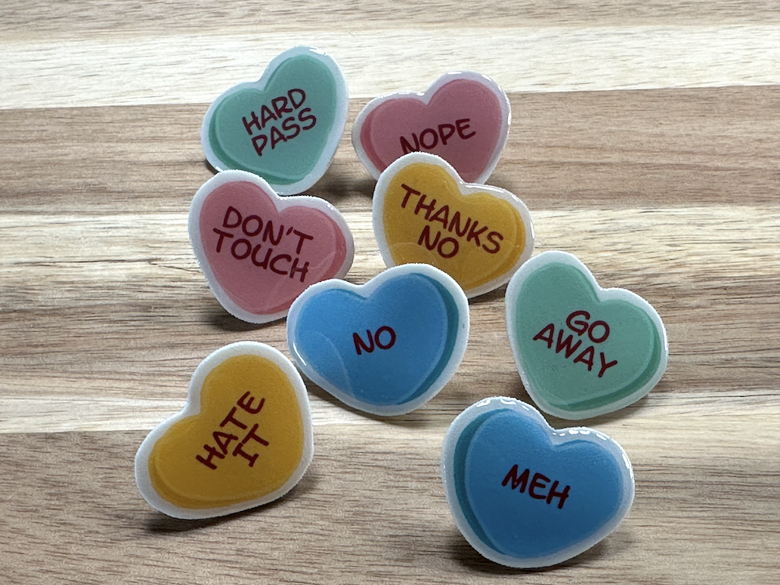 Colorful heart-shaped stickers with messages such as 'HARD PASS', 'NOPE', 'DON'T TOUCH', 'THANKS NO', 'GO AWAY', 'HATE IT', 'NO', and 'MEH' placed on a wooden surface.