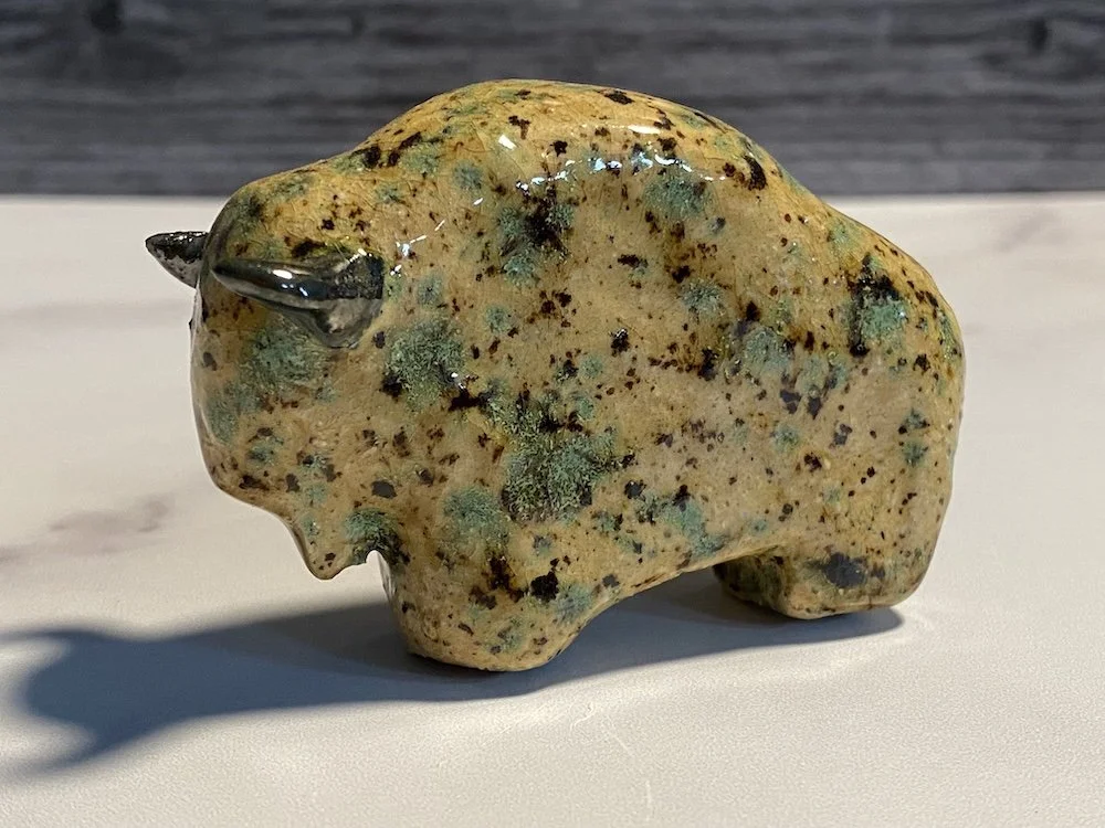 A small, handmade ceramic sculpture of a bear, with a yellowish glaze and green and black speckles, and black horns on its head, placed on a white surface with a shadow cast beside it.