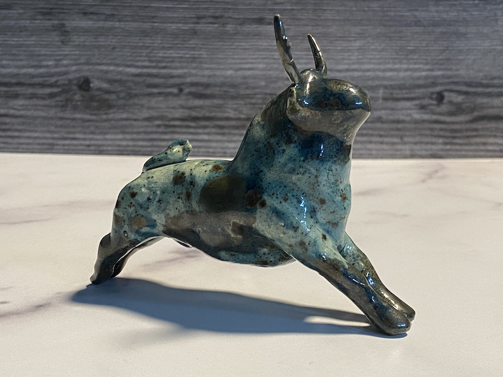 Ceramic sculpture of a stylized dog with elongated ears, painted in a blue and brown mottled glaze, standing on all fours on a white surface against a gray wood-paneled background.