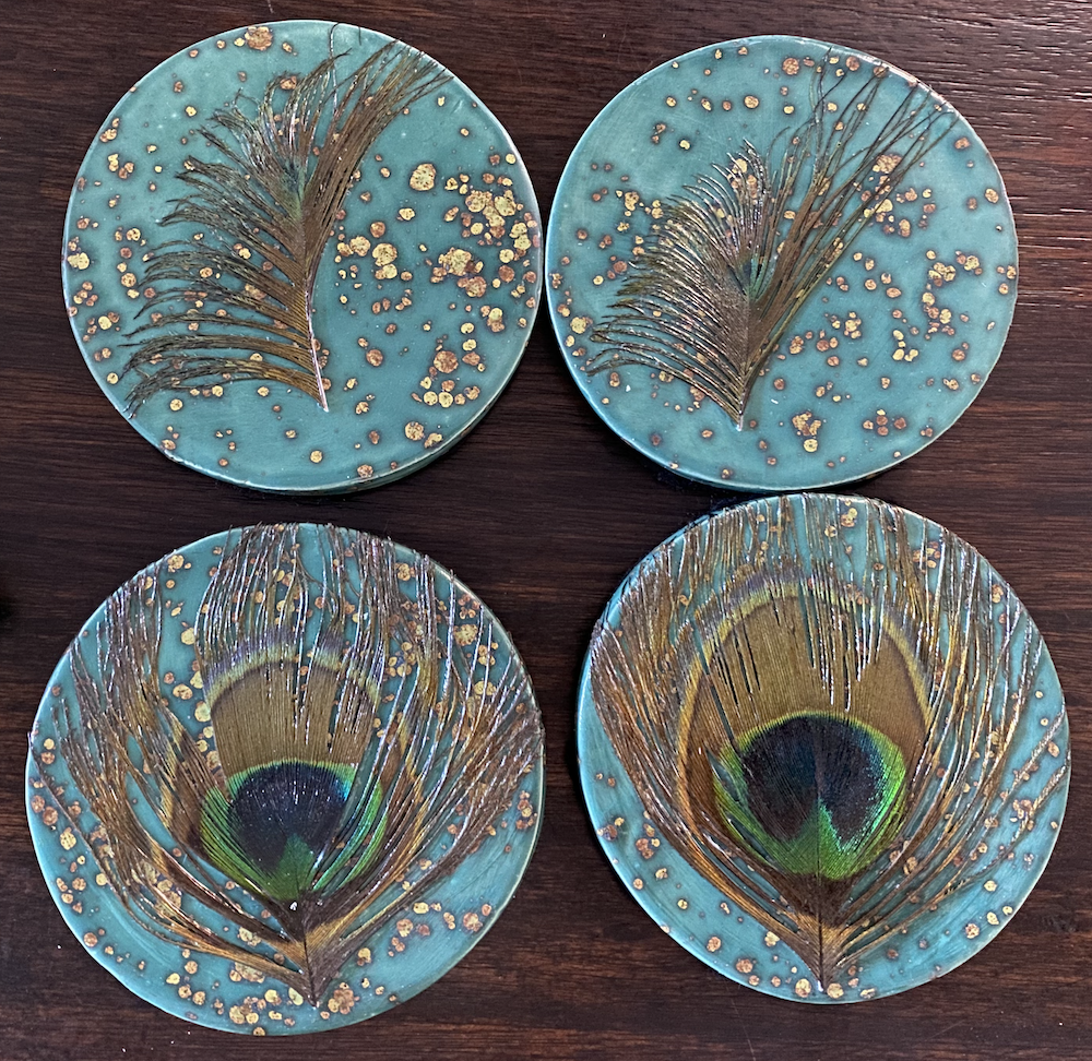 Four decorative plates featuring peacock feather designs with a turquoise background and gold accents, arranged on a dark wooden surface.