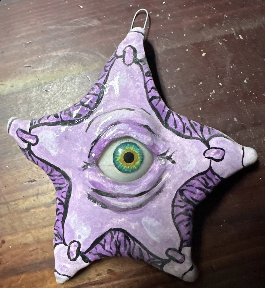 A star-shaped decorative object with a large eye in the center, painted purple with black and white details, placed on a wooden surface.