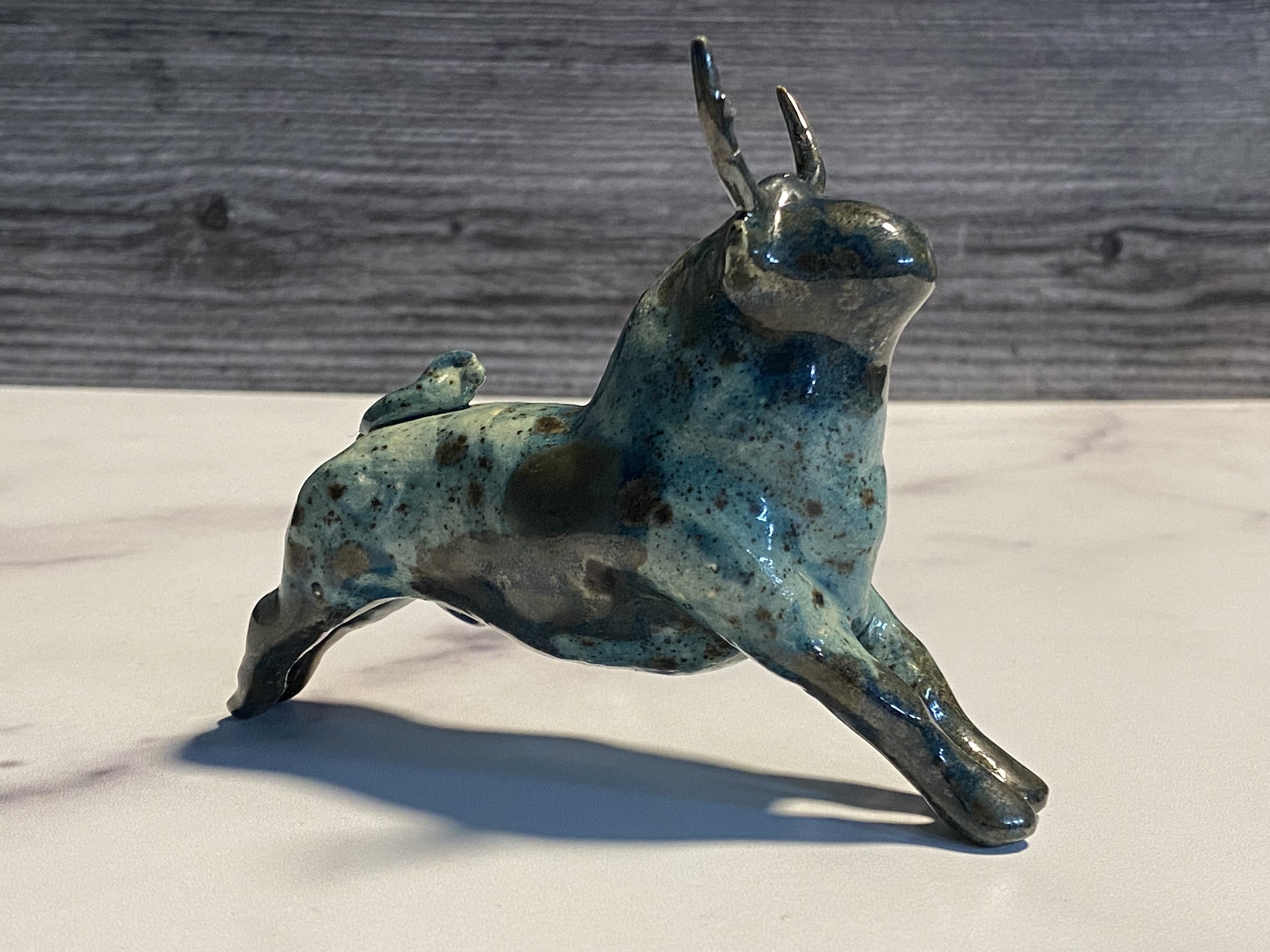 A small, abstract sculpture resembling a rabbit with a metallic blue and bronze finish. The sculpture has elongated ears and a curved body, positioned on a white surface with a wooden-textured background.