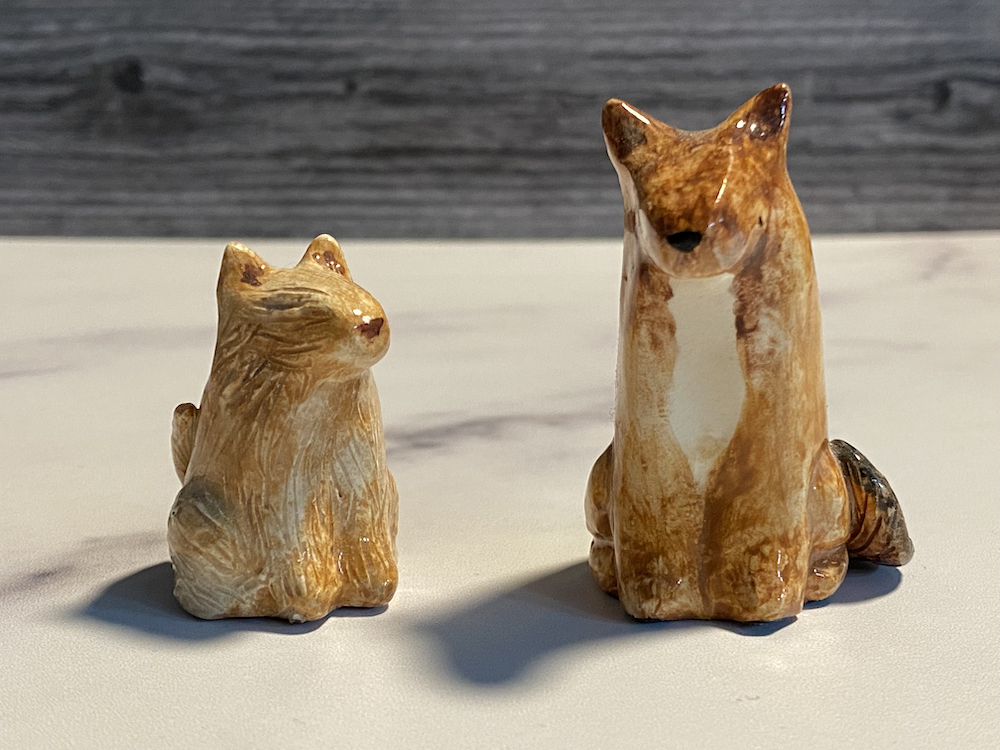 Two ceramic fox figurines on a white surface with a dark wooden background.