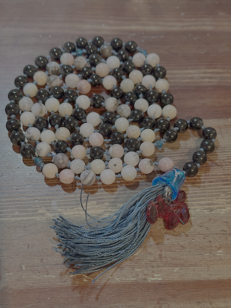 A string of prayer beads or rosary with black, white, and marbled beads, a blue section near the tassel, red gemstone accents, and a gray tassel, resting on a wooden surface.
