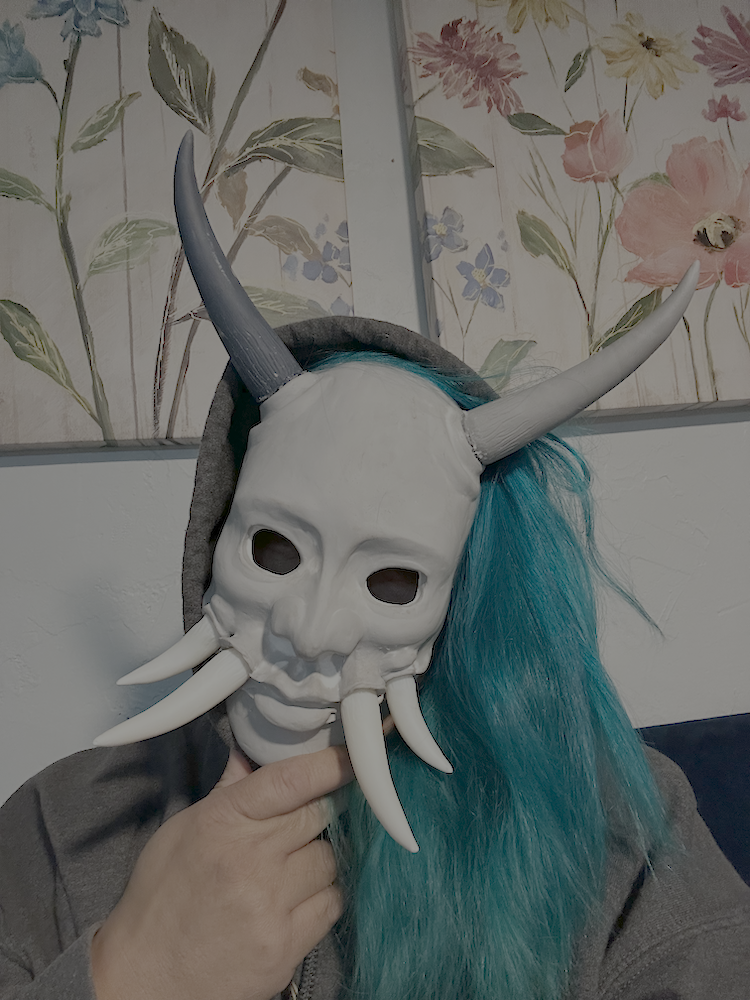 A person wearing a white oni skull mask with large horns and tusks, and a blue wig, sitting indoors with floral artwork in the background.
