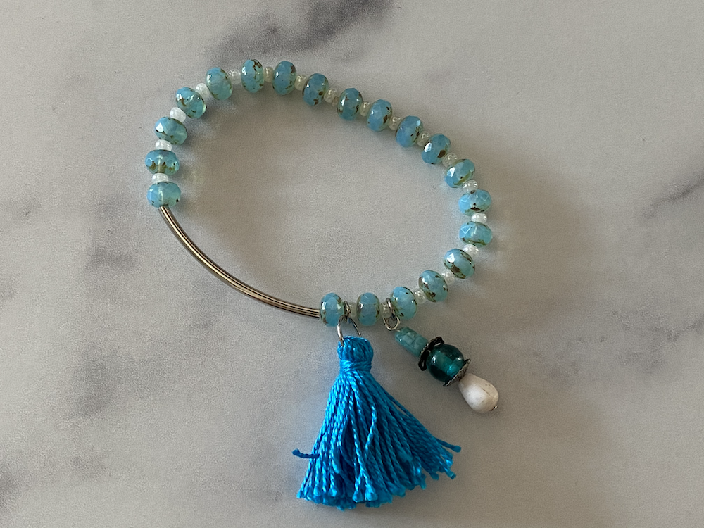 Light blue beaded bracelet with a blue tassel and decorative charms, placed on a light gray surface.