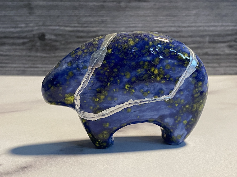 A carved stone shaped like a bear with a blue and yellow speckled pattern resting on a white surface with a wooden background.