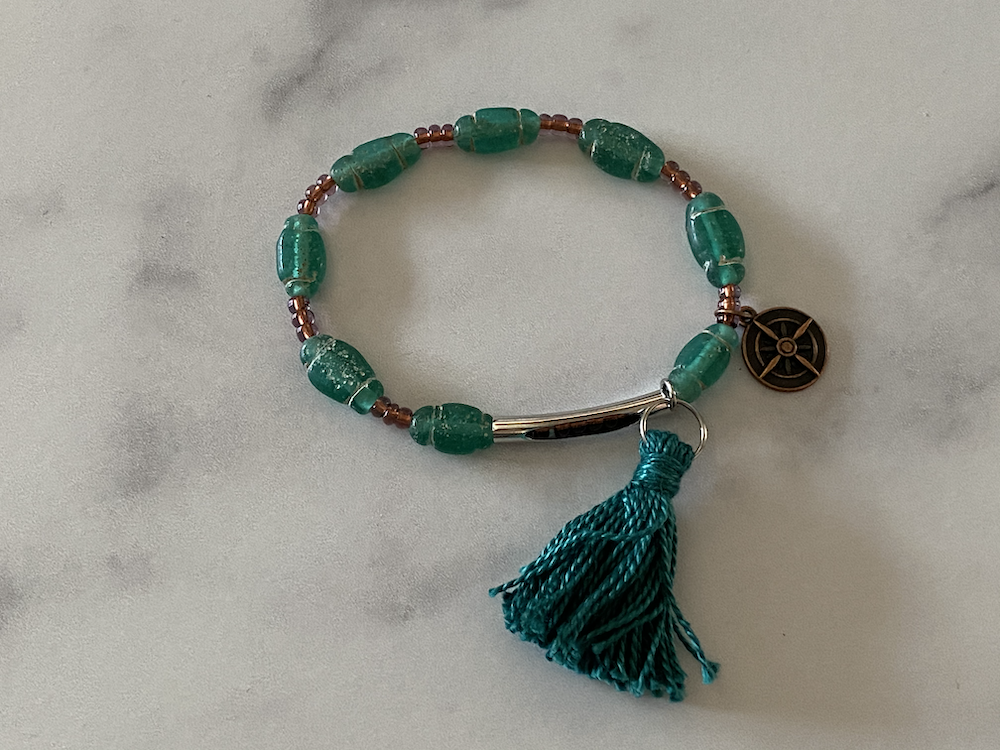 Beaded bracelet with green and brown beads, a silver metal clasp, a black round charm, and a teal tassel.