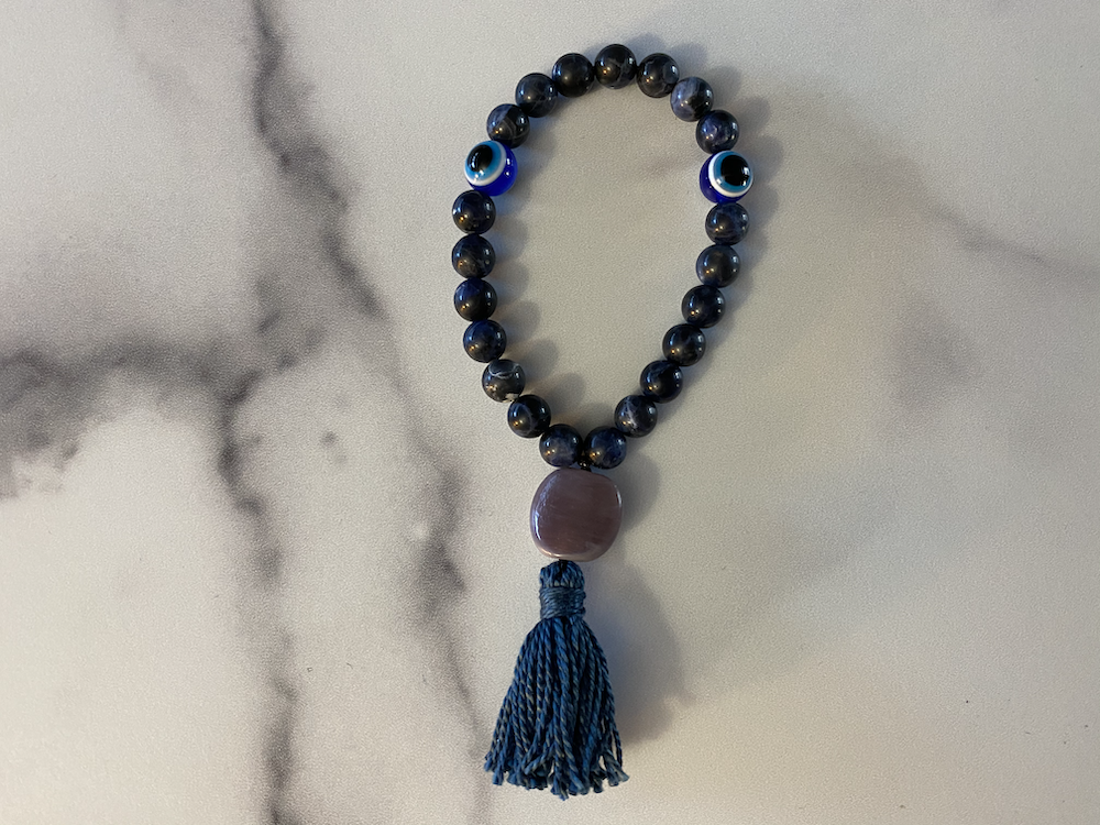 Beaded bracelet with dark round beads, two eye-shaped beads, a large pink bead, and a blue tassel, resting on a light-colored marble surface.