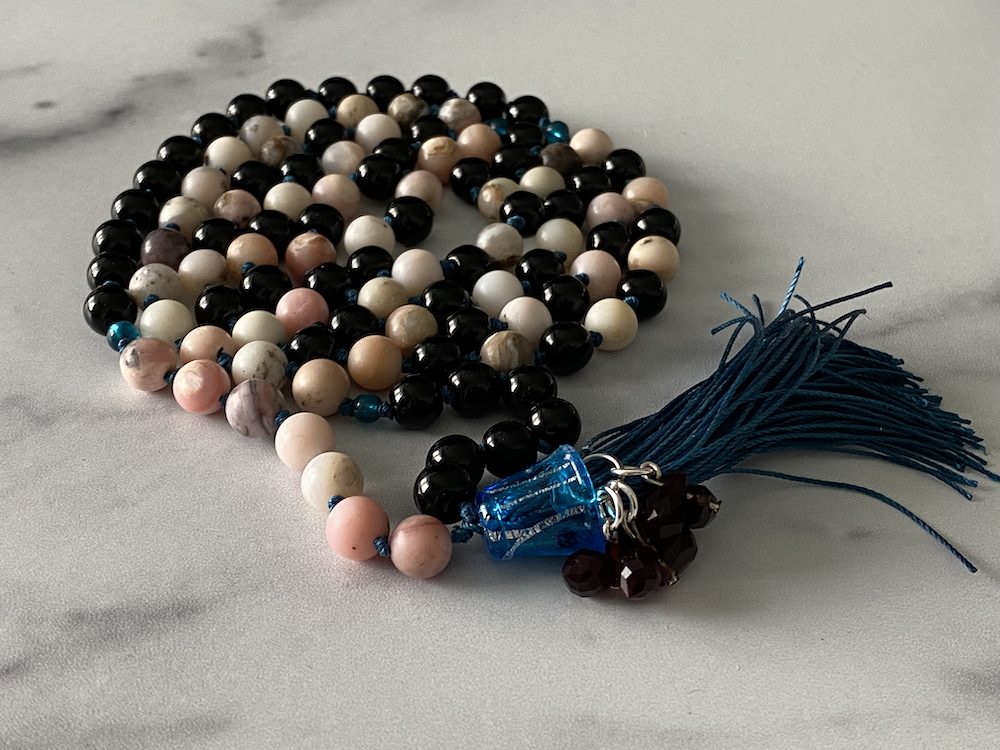 Beaded necklace with black, white, pink, and marbled beads, blue glass charms, and a blue tassel, lying on a light grey surface.