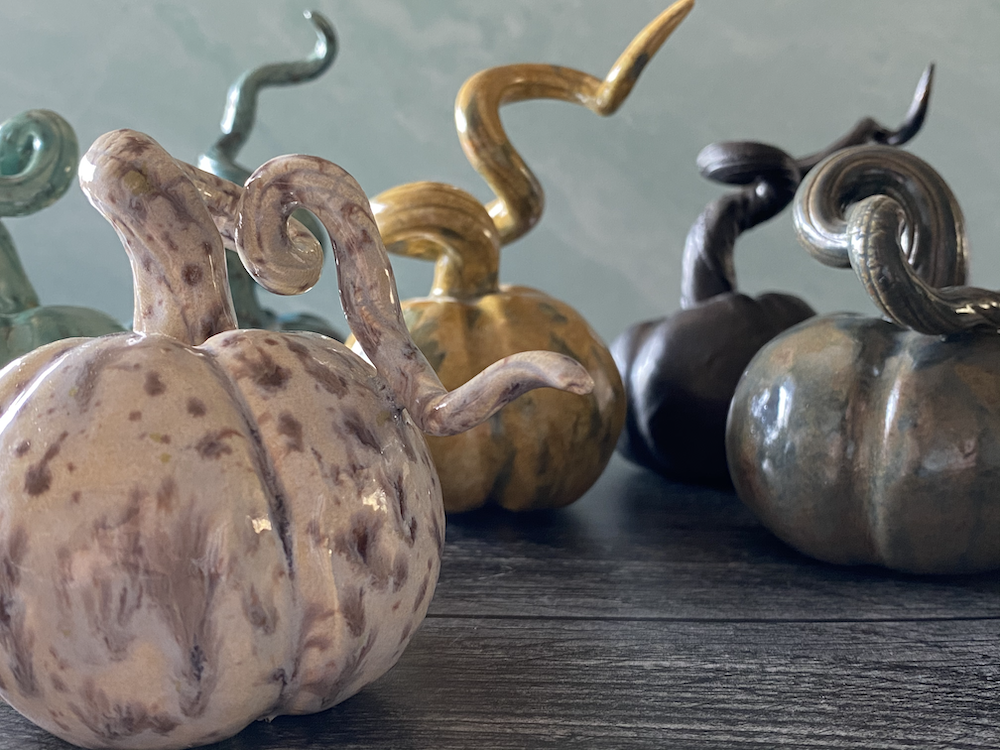 Four decorative glass pumpkins with curly stems in pastel, yellow, black, and green colors, placed on a dark wooden surface.