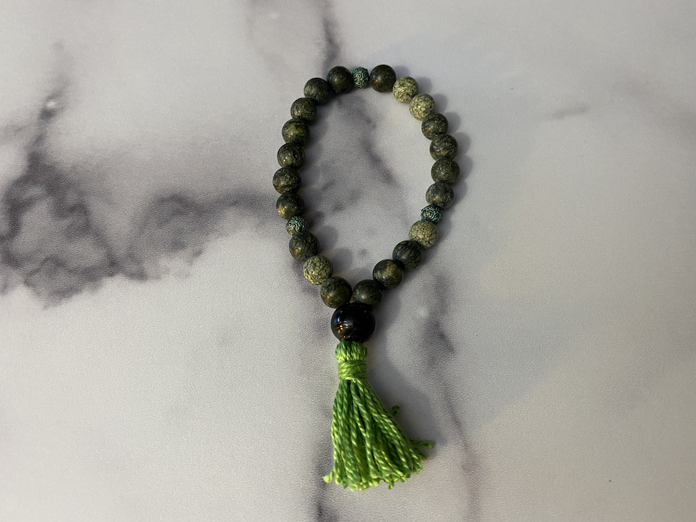 A beaded bracelet with green and black beads, featuring multiple small green beads, a large black bead, and a bright green tassel hanging from it, placed on a light gray marble surface.