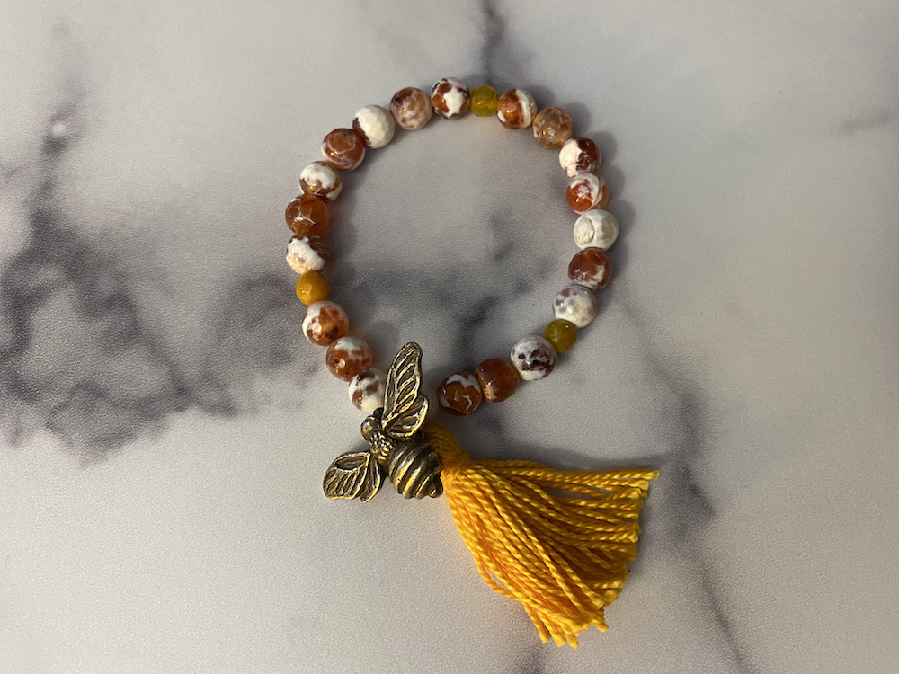 Beaded bracelet with a bee charm and a yellow tassel on a gray and white marble surface.