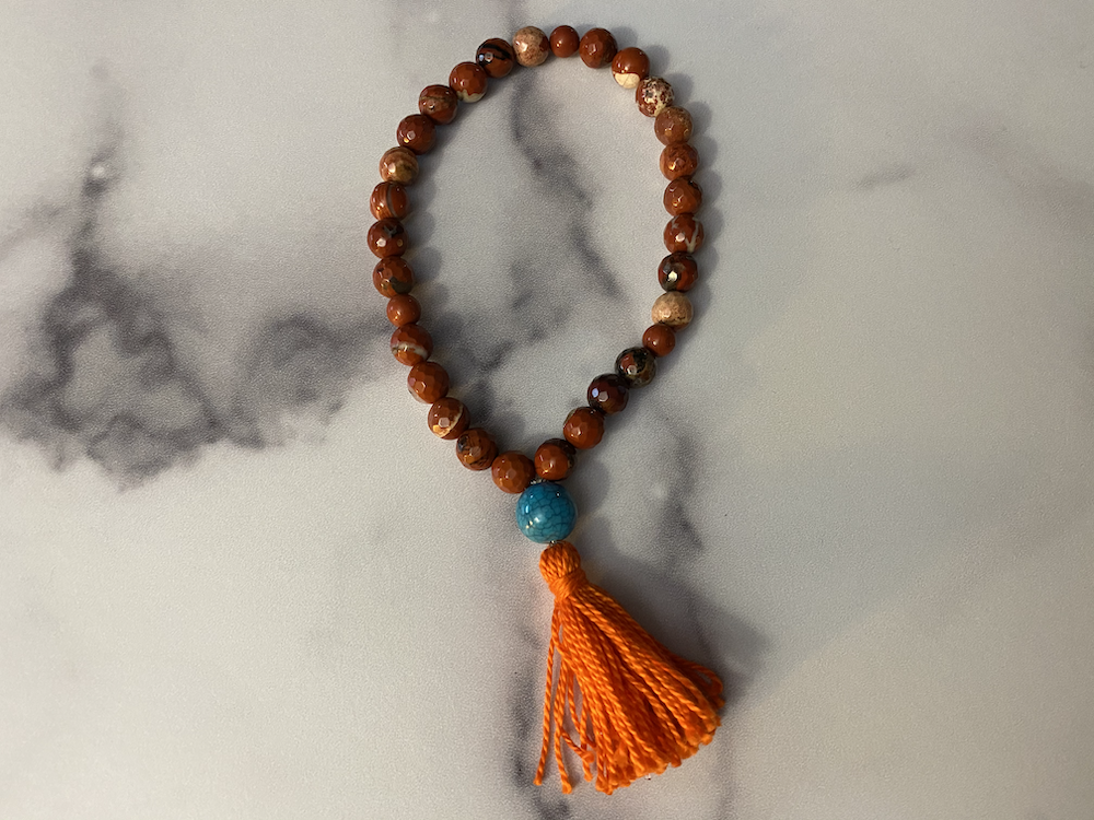 Beaded prayer necklace with a blue bead at the center and an orange tassel at the bottom, lying on a marble surface.