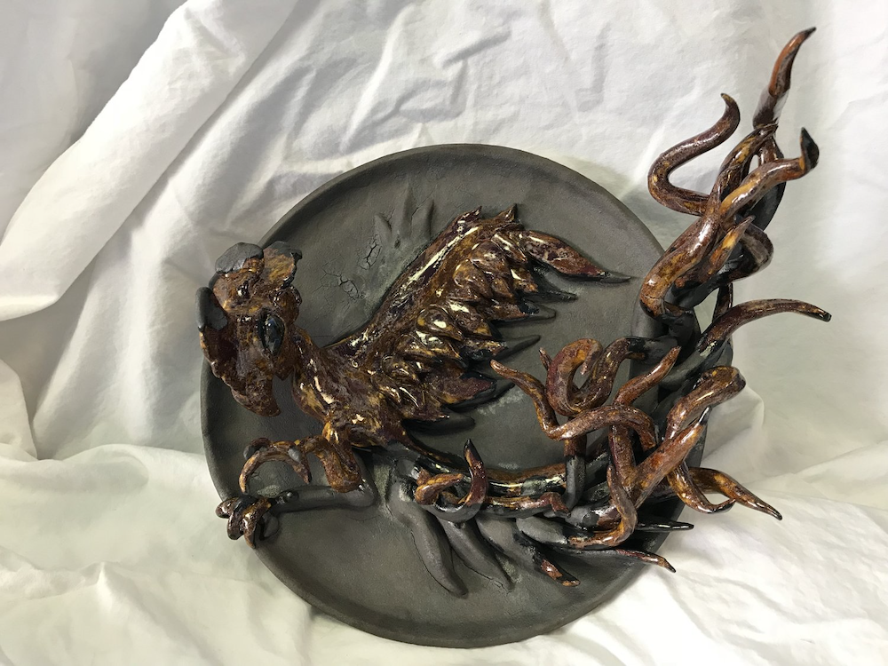 A metal sculpture of a dragon with wings, claws, and a tail, mounted on a round black base, displayed on a white fabric background.