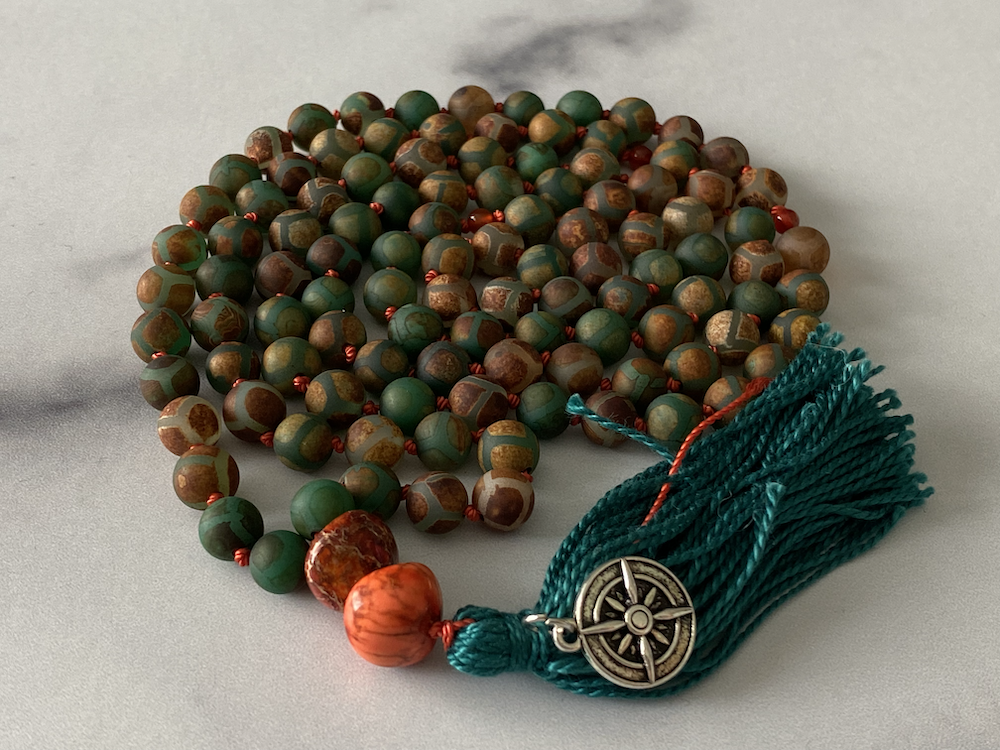 A string of prayer beads, primarily green with some brown and orange beads, include a decorative tassel and a metal charm with a compass design.