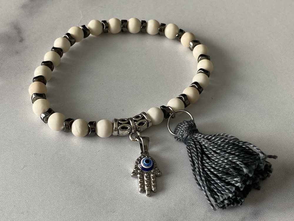 Beaded bracelet with black and white beads, a silver hand-shaped charm with an eye, and a black and white tassel, on a gray surface.