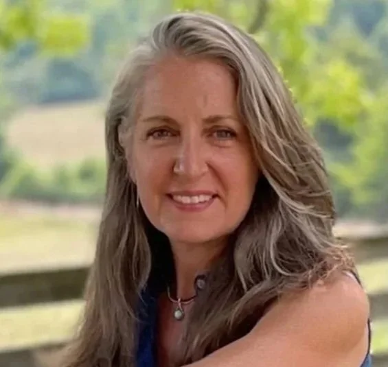 A woman with long gray hair smiling outdoors in a natural setting with trees and a fence in the background.