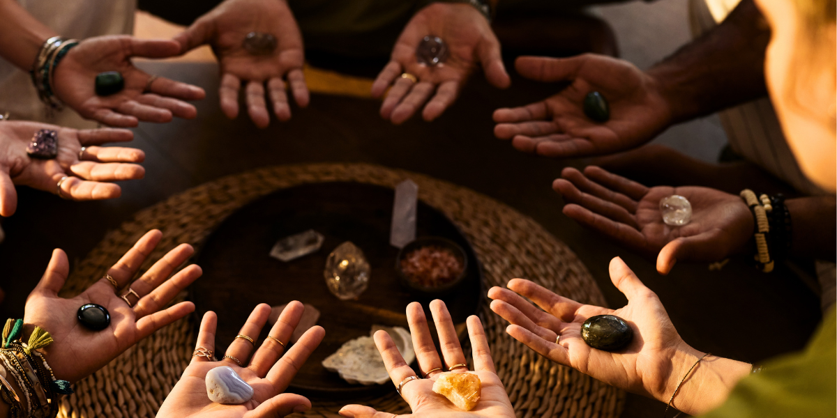 Multiple people with various rings on their fingers holding polished stones and crystals, gathered around a table with a display of stones and crystals.