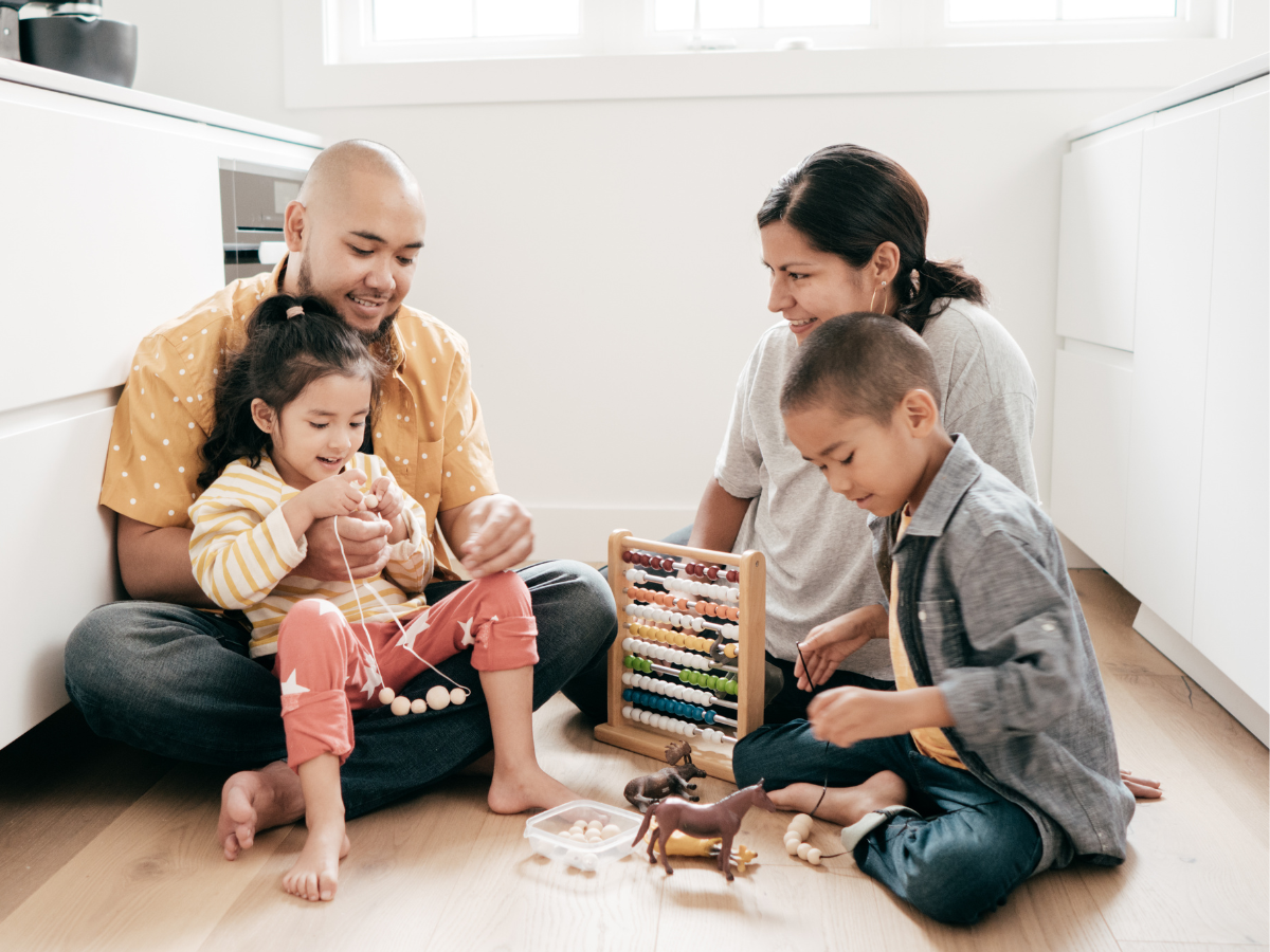 A family of four playing with toys and a bead abacus on the floor of a bright kitchen.